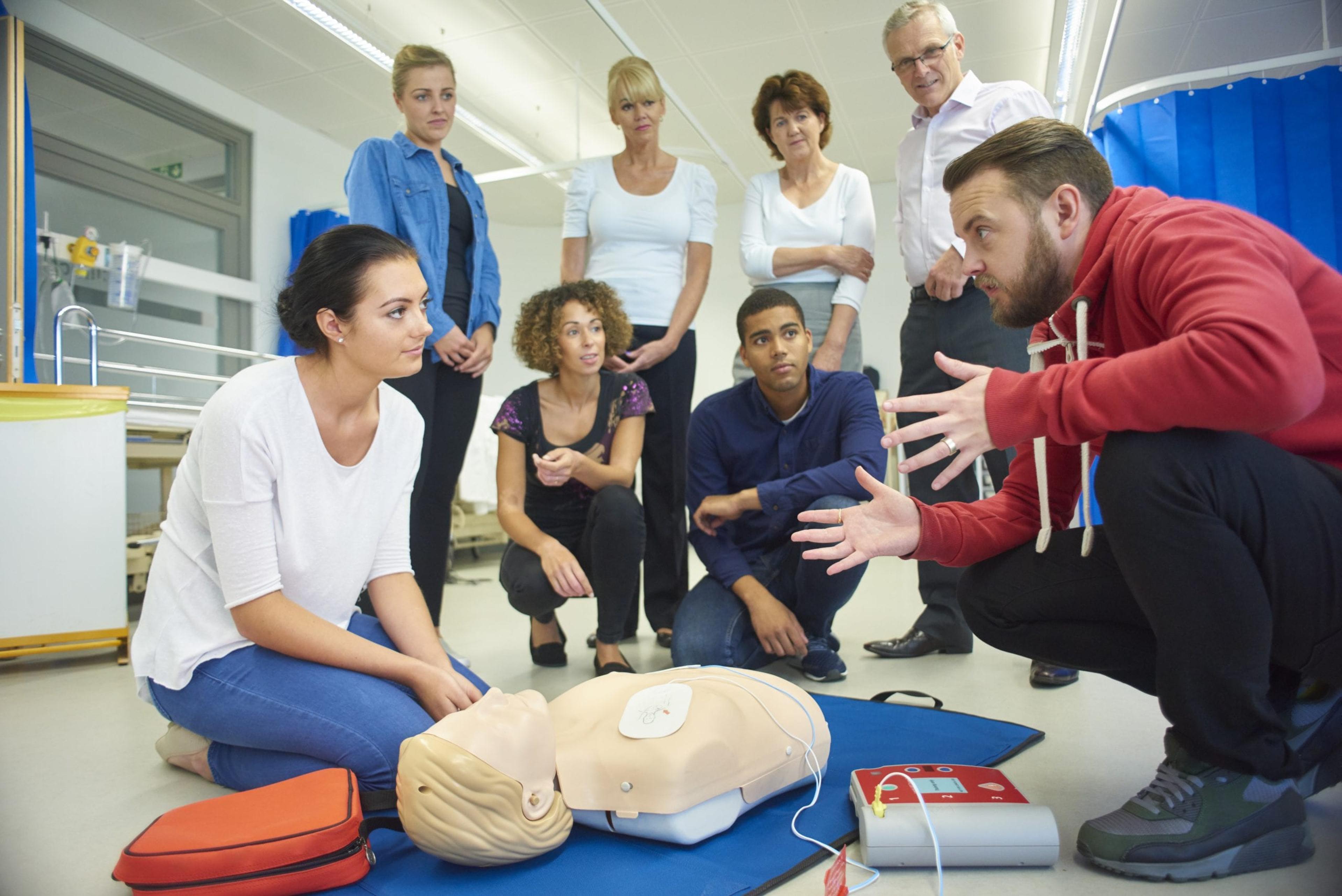 a mixed age group listen to their tutor as he shows the procedure involved to resuscitate using a defibrillator.