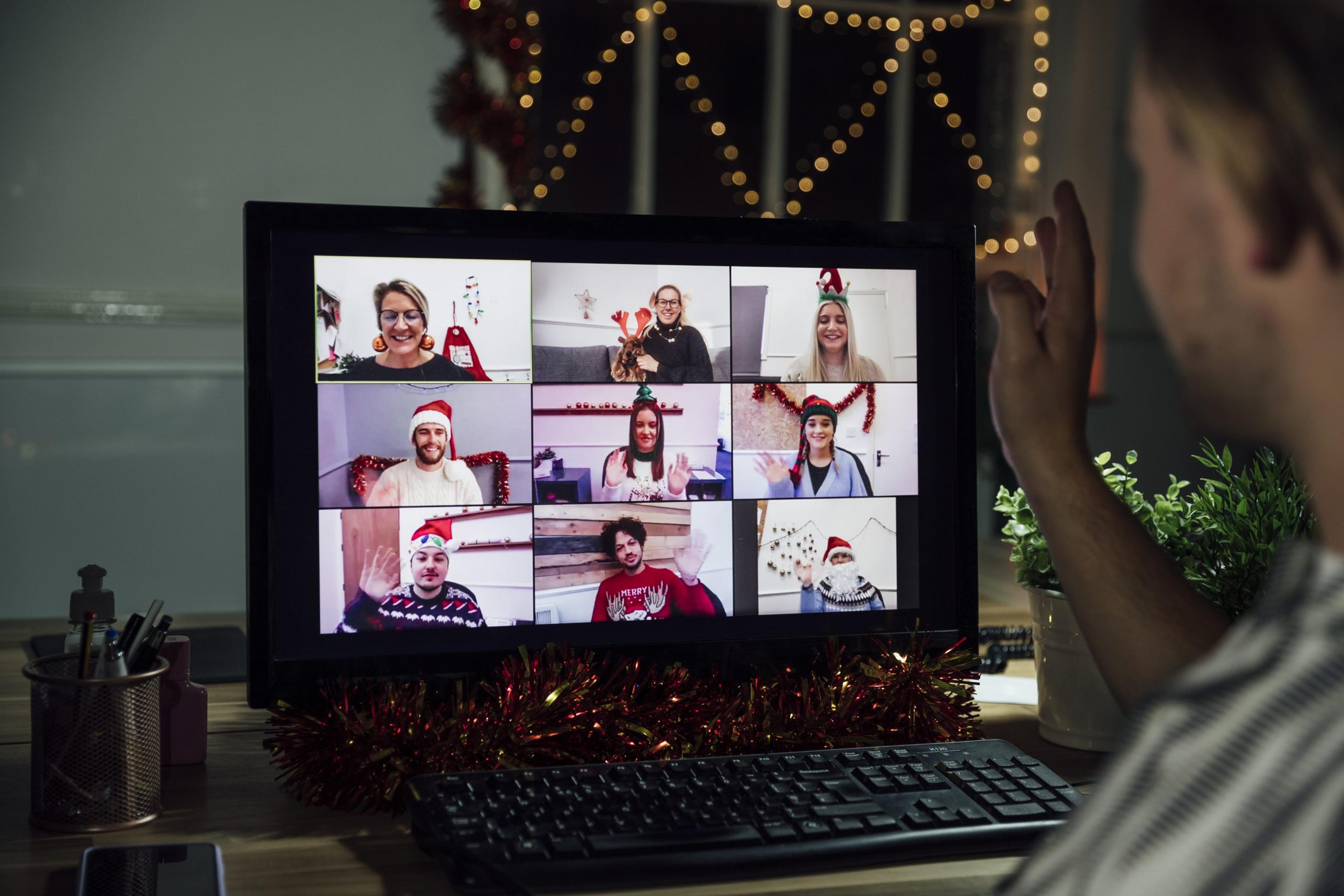 Over the shoulder view of a festive group of colleagues having an office party via video call. They are all greeting each other and chatting whilst wearing festive clothing. A man is sitting at his computer having to the screen.