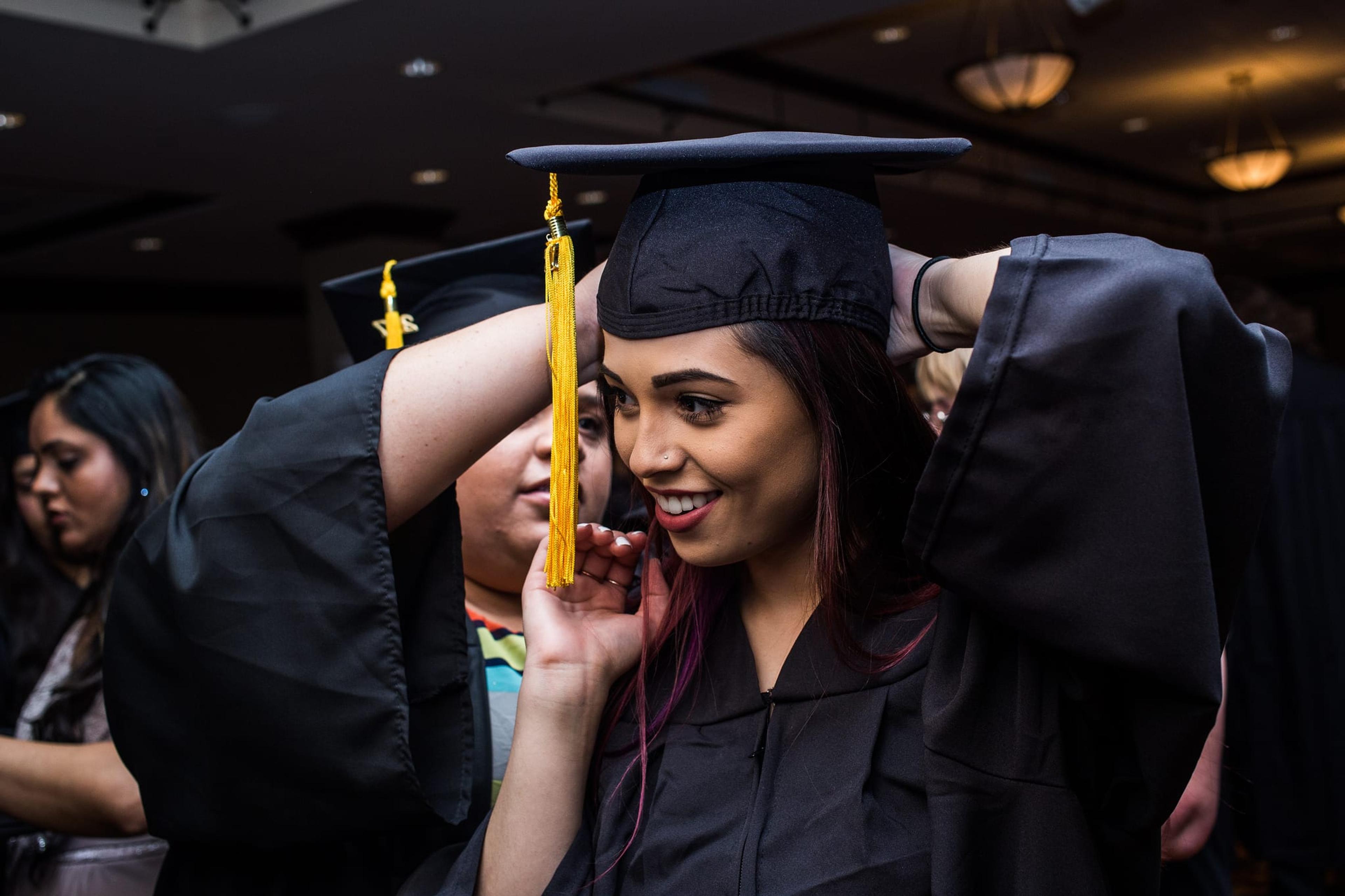 Image of a woman about to graduate from college in cap and gown. A friend is adjusting her cap for her.