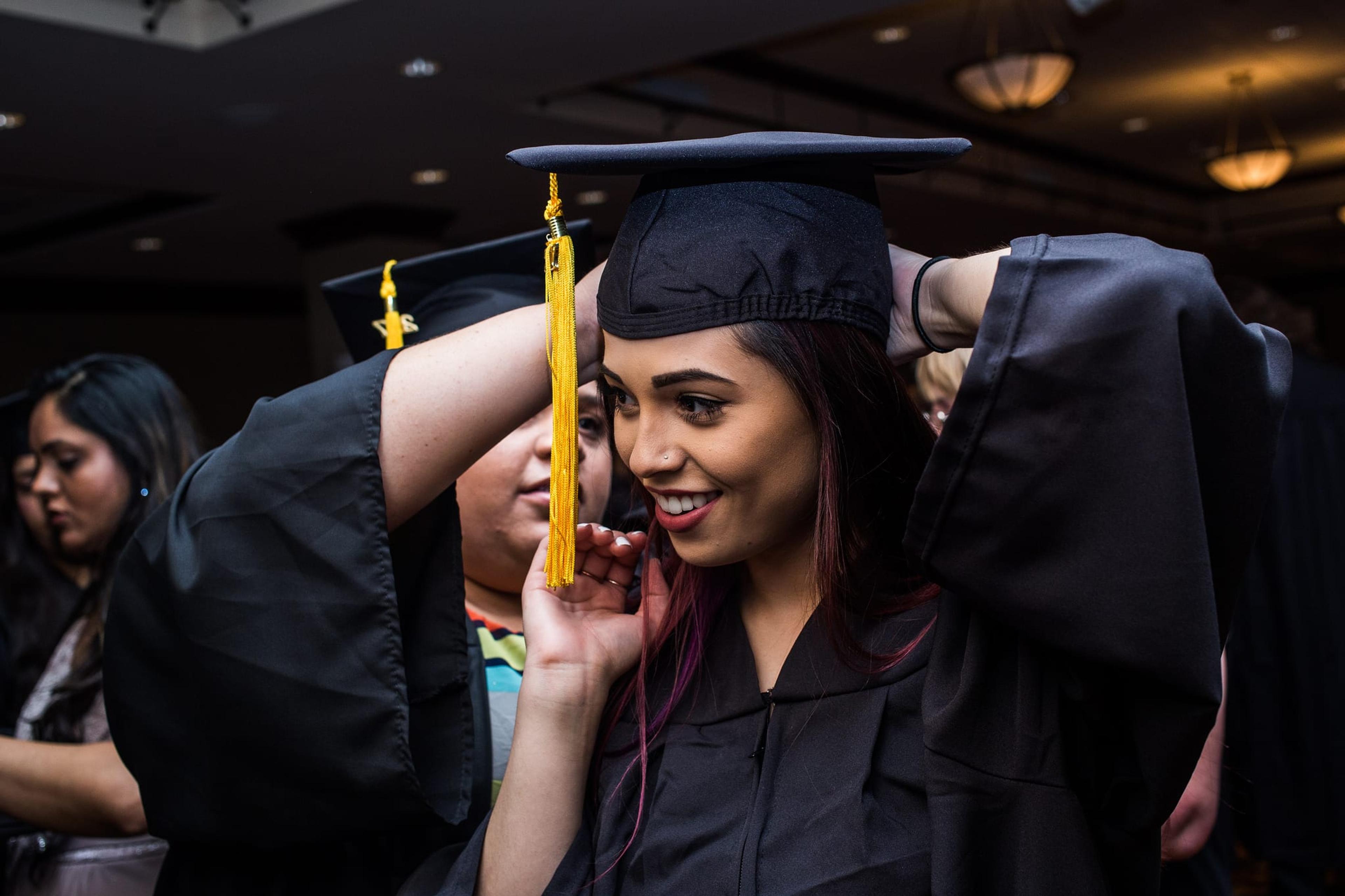 Image of a woman about to graduate from college in cap and gown. A friend is adjusting her cap for her.