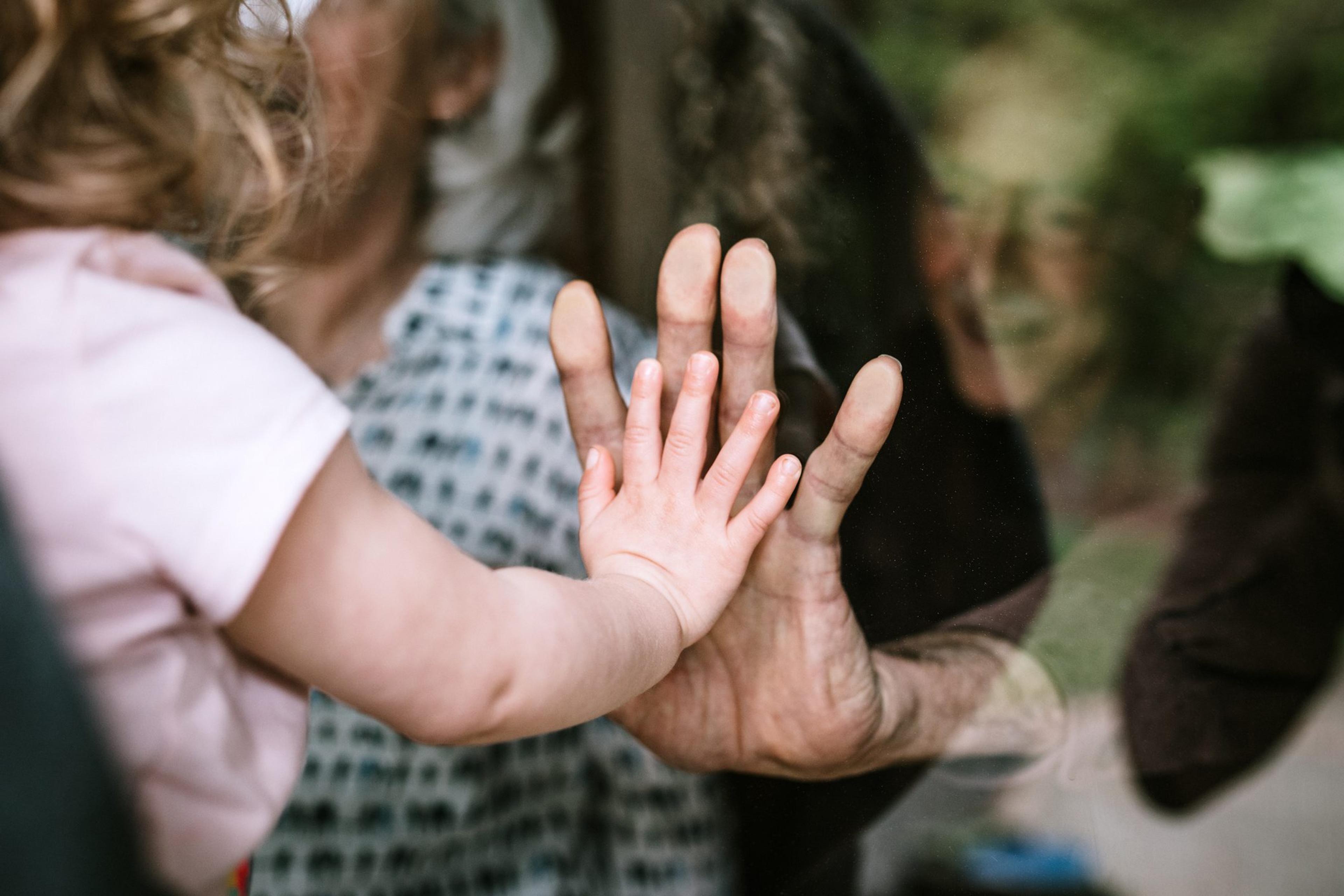 A mother stands with her daughter, visiting senior parents but observing social distancing with a glass door between them. The granddaughter puts her hand up to the glass, the grandfather and grandmother doing the same. A small connection in a time of separation during the Covid-19 pandemic.