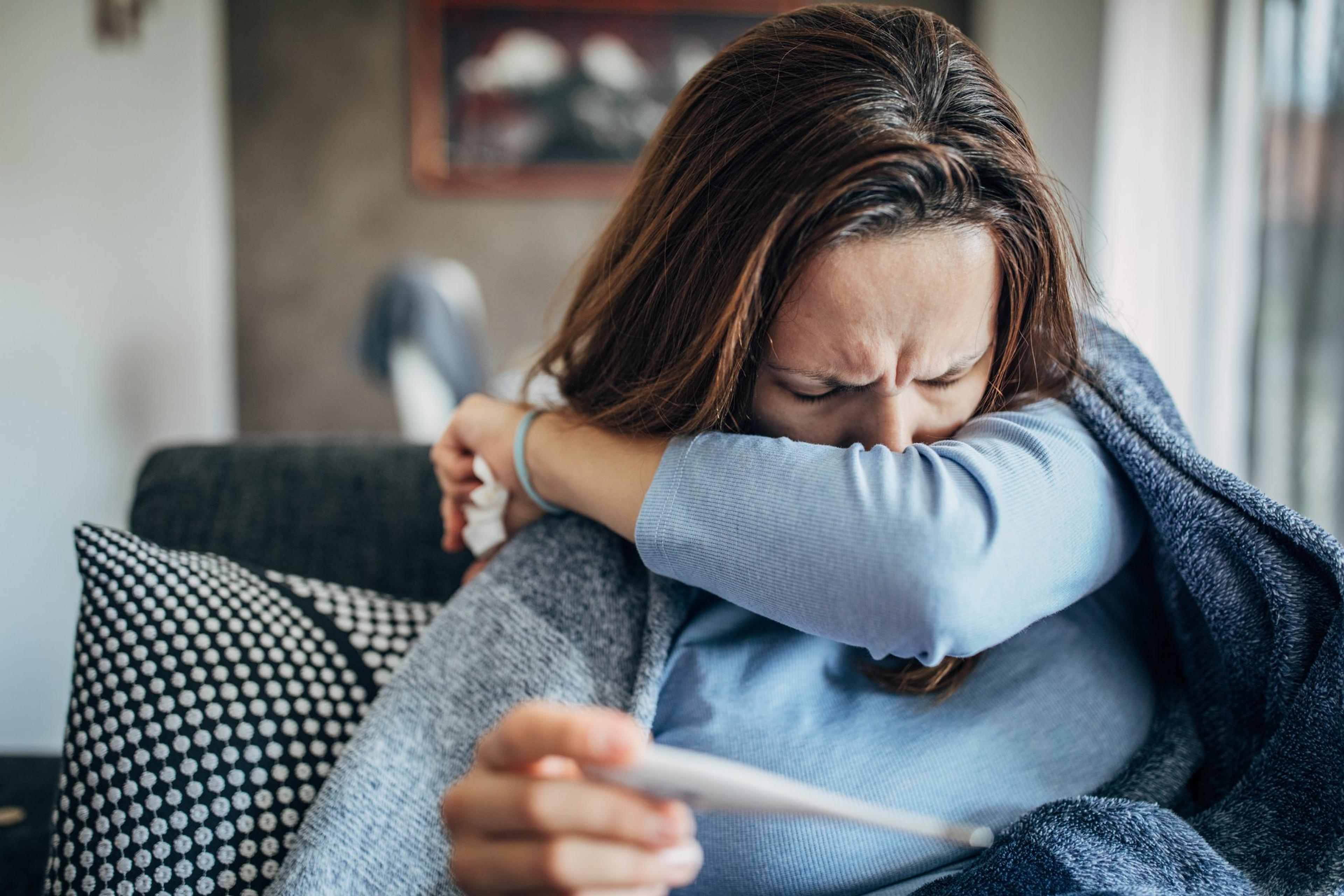 Woman with fever symptoms sitting on sofa and holding thermometer