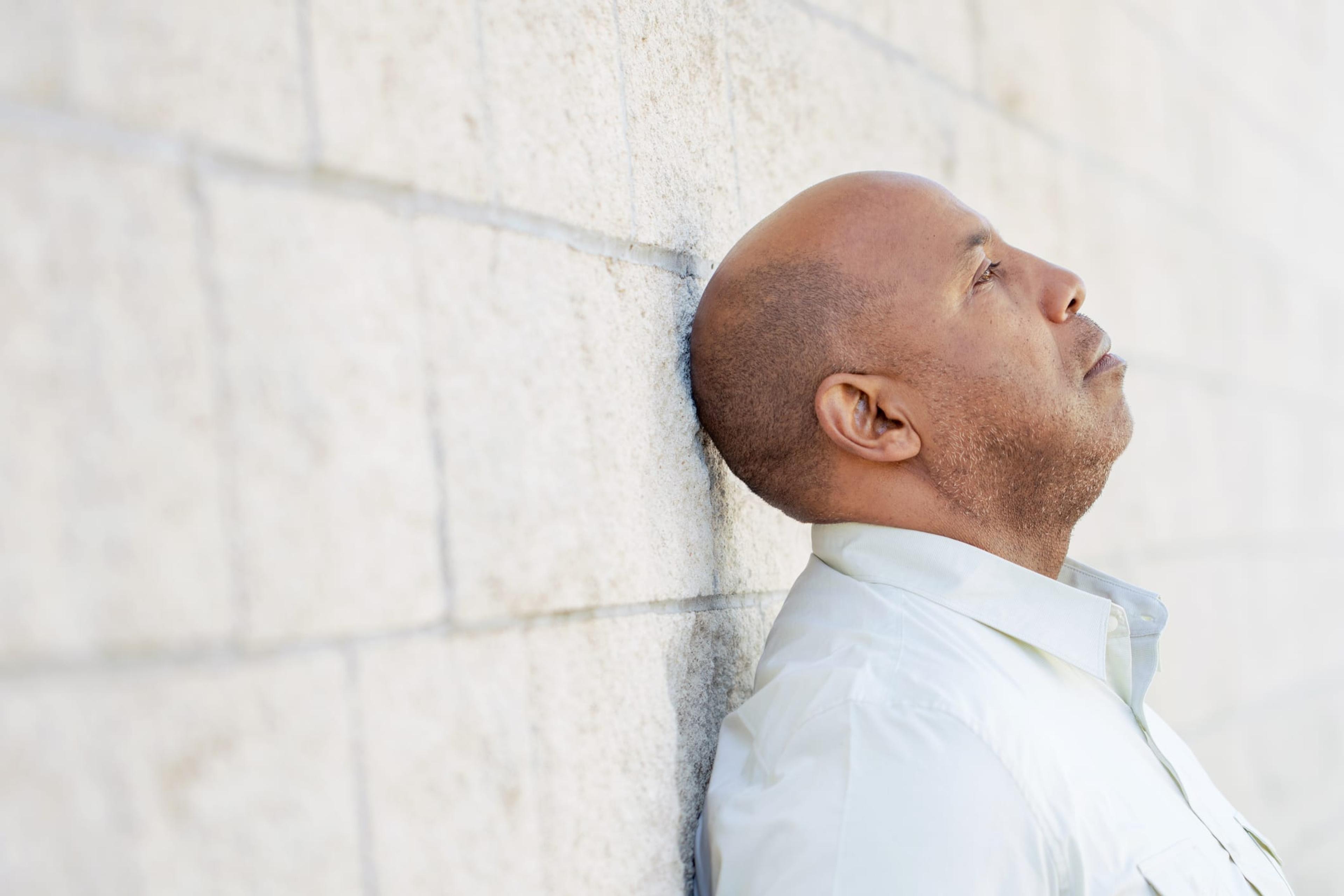 Man in work shirt leaning up against wall, contemplative look on his face.