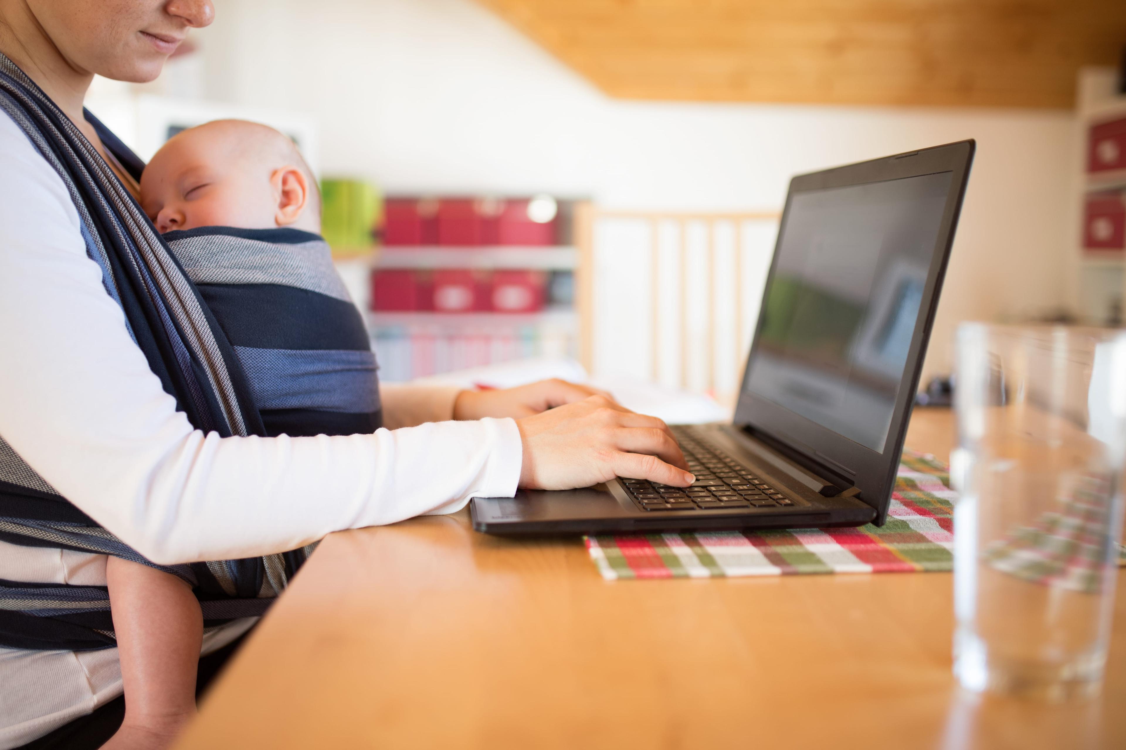 A woman with a baby in a baby carrier working on a laptop.
