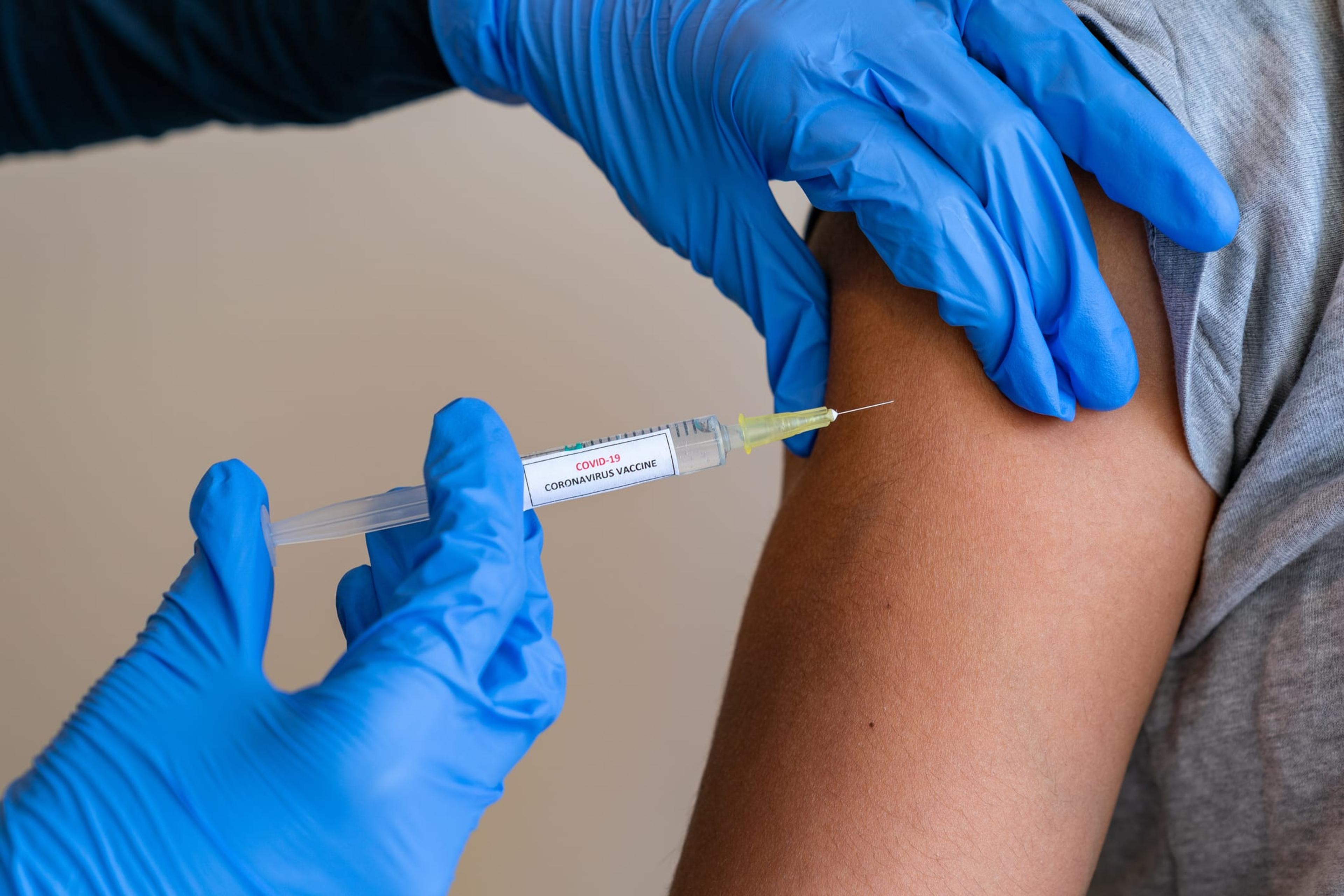Female doctor wearing blue latex gloves injecting female with a needle and syringe containing a dose of the COVID-19 vaccine.