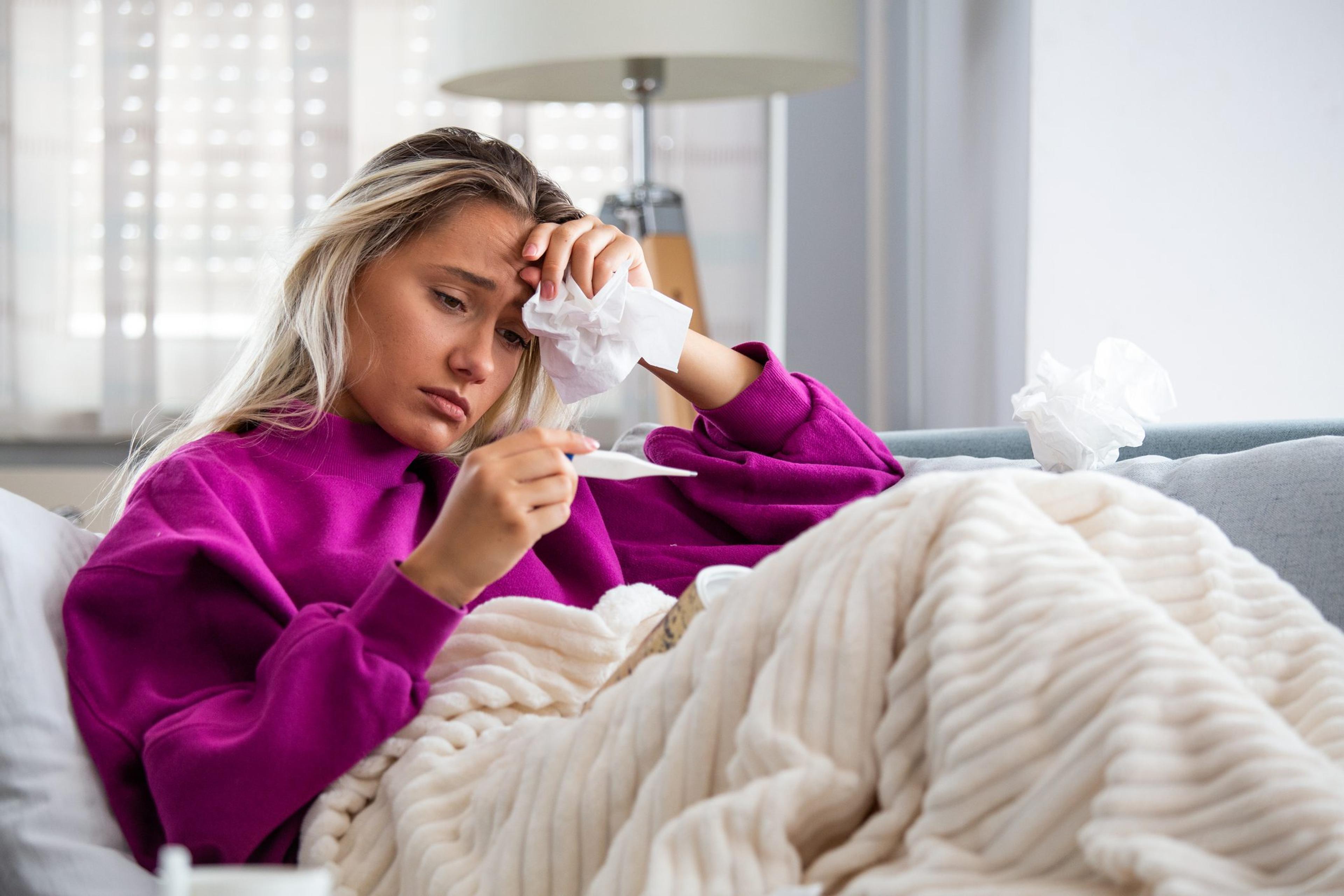 Young woman sick with the flu sitting on the couch at home looking at a thermometer