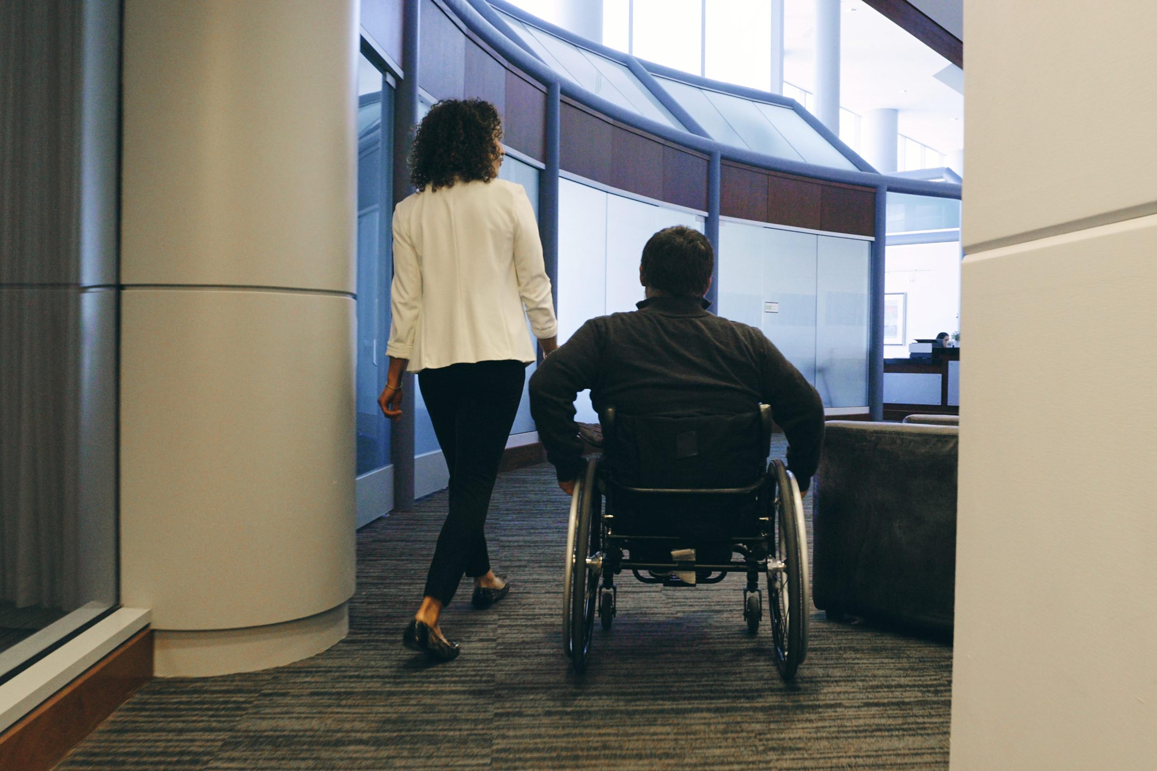 A person walks next to another person in a wheelchair in an office