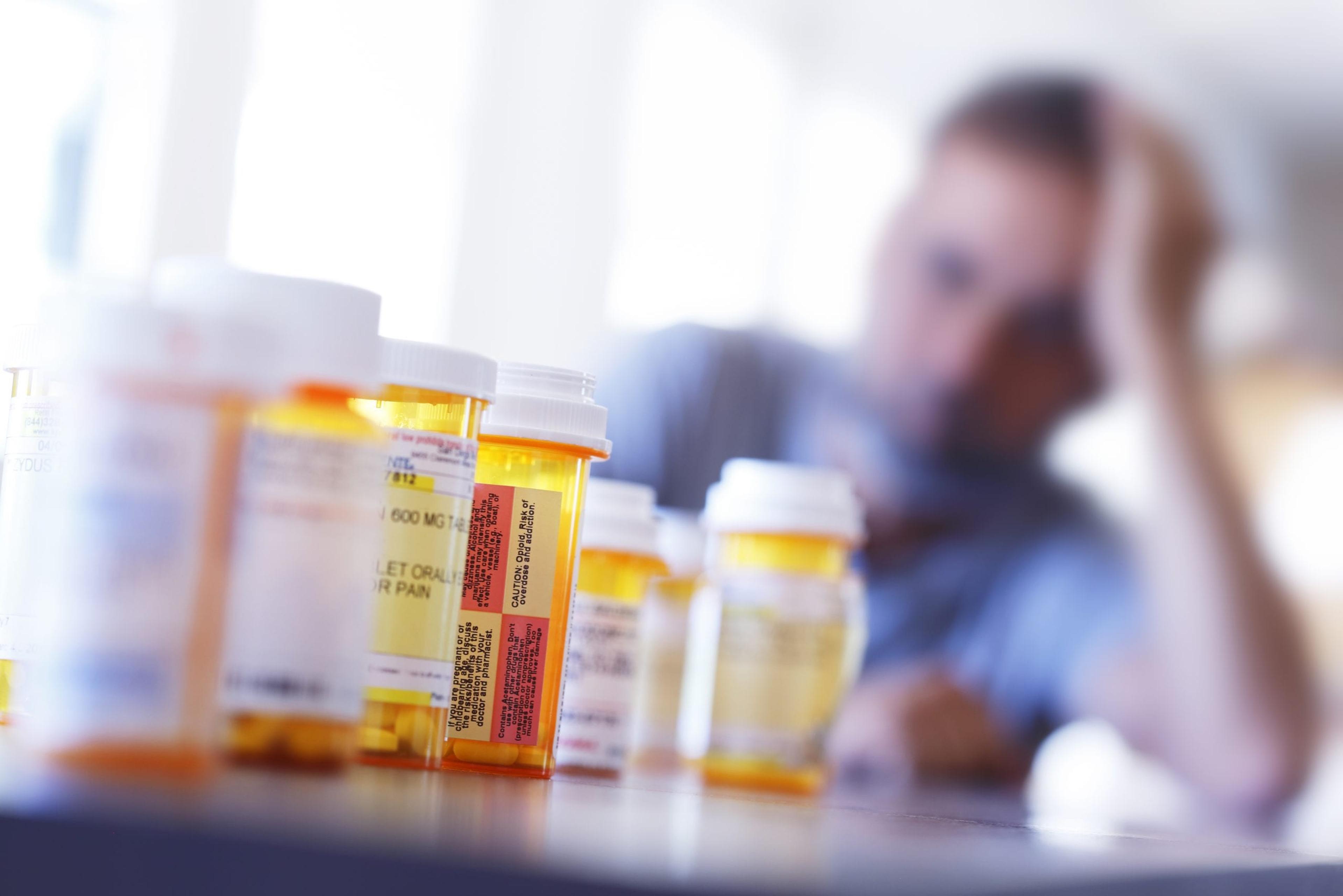 Large group of prescription bottles in the foreground with man sitting at table in background.