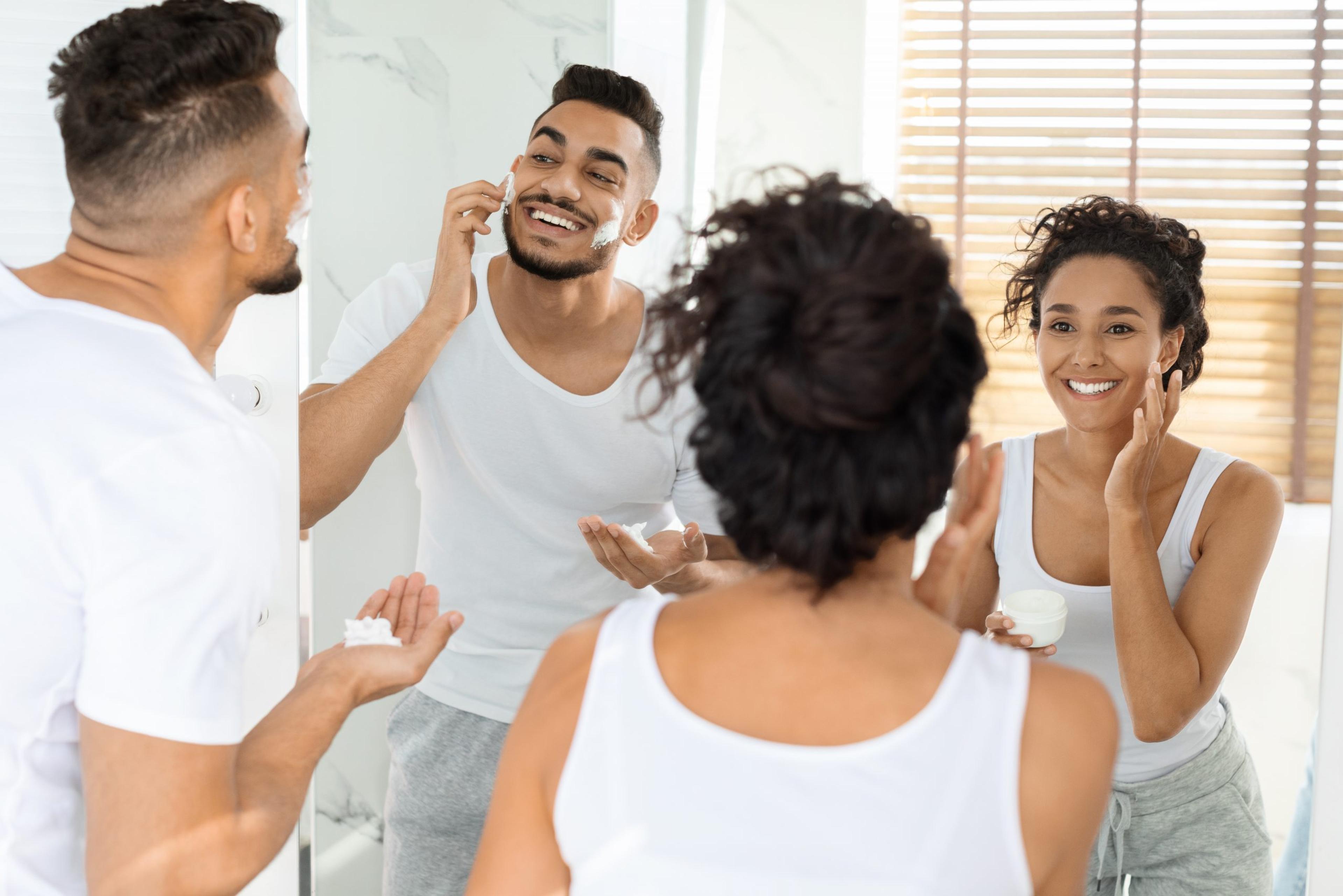 Morning Routine. Happy Young Arab Couple Getting Ready Together Near Mirror In Bathroom, Woman Applying Moisturising Cream While Man Using Shave Foam, Selective Focus On Reflection, Free Space