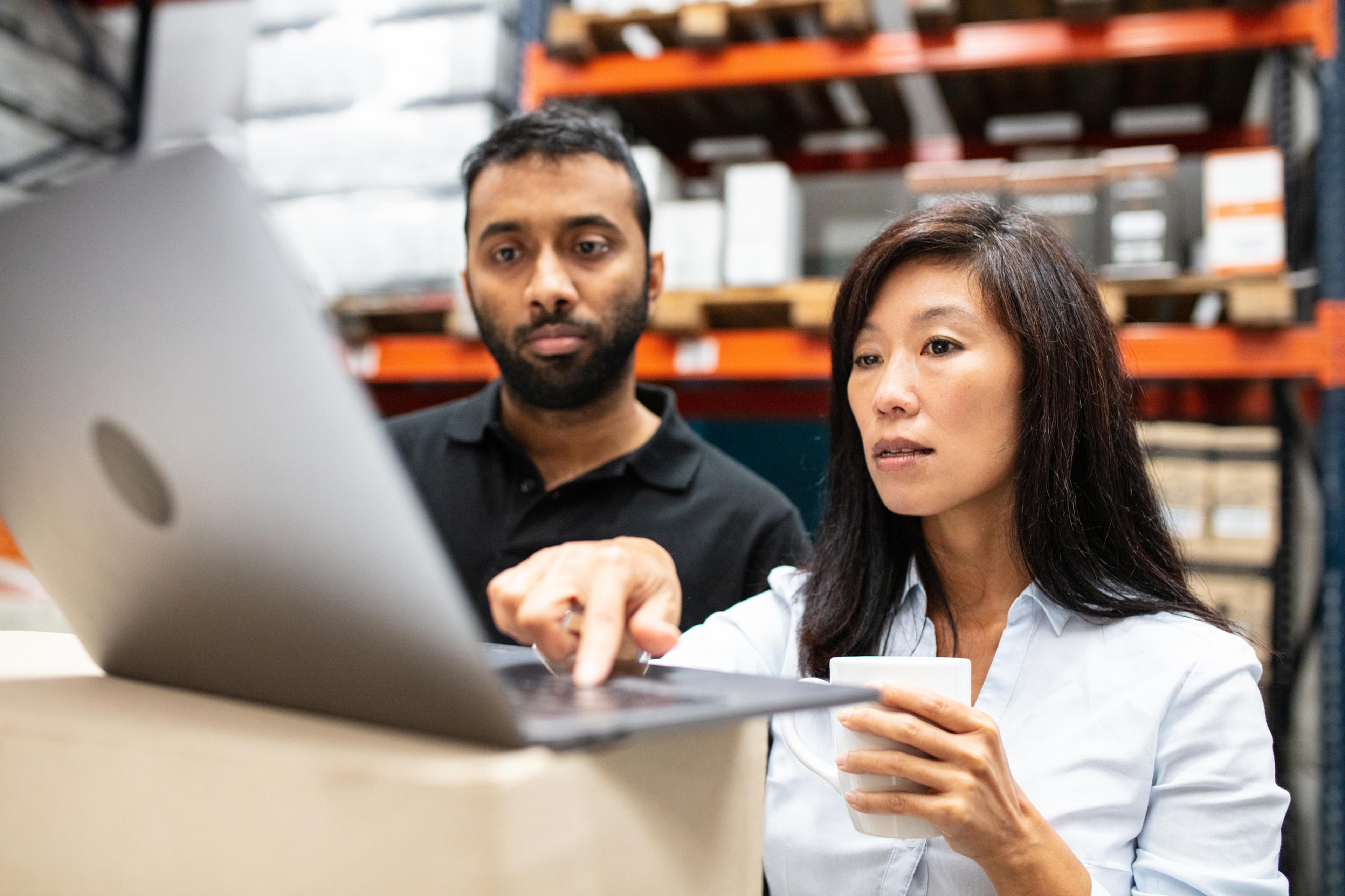 A woman and a man look at a laptop screen in a warehouse.