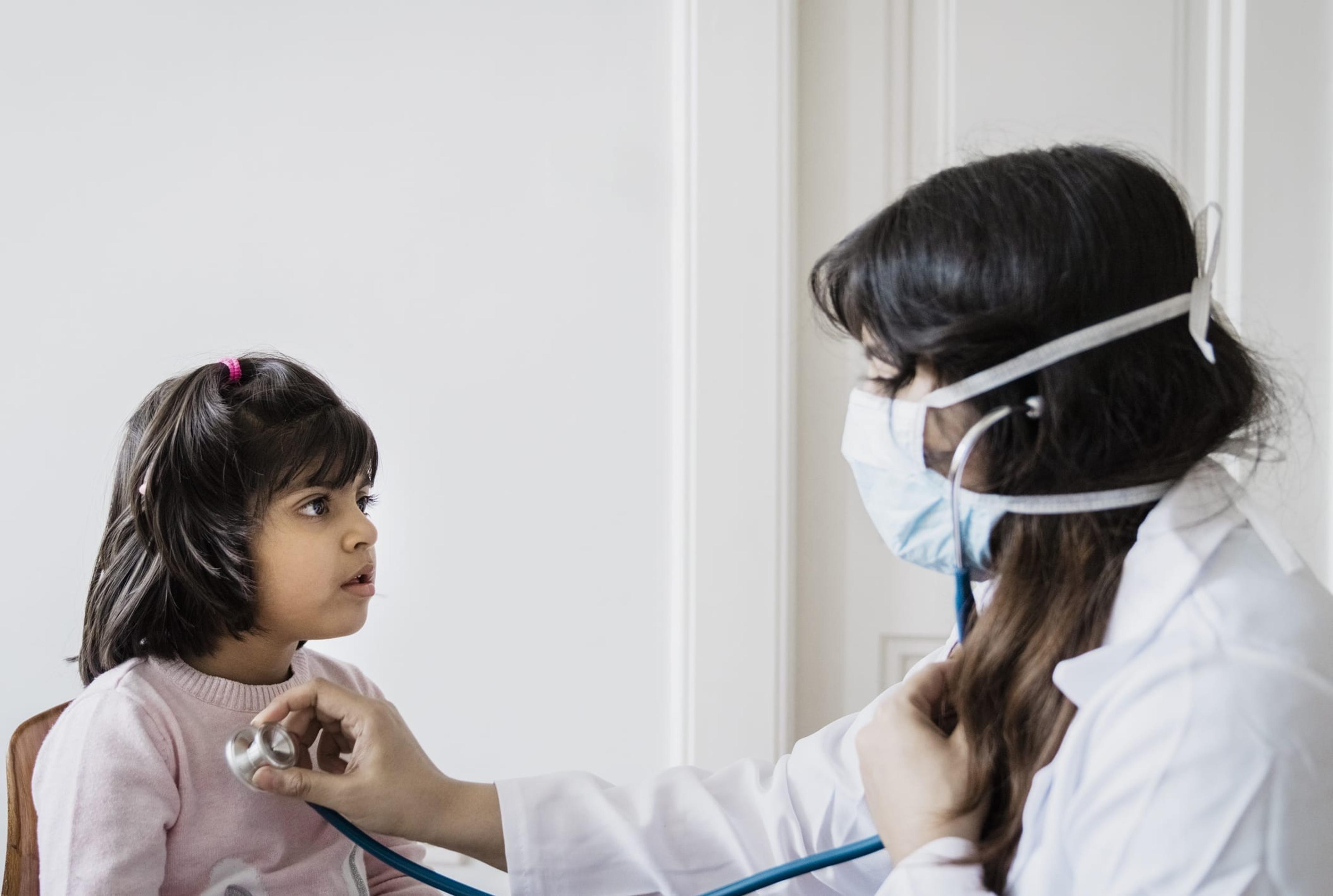 Female doctor wearing a mask listens to the heartbeat of a young girl