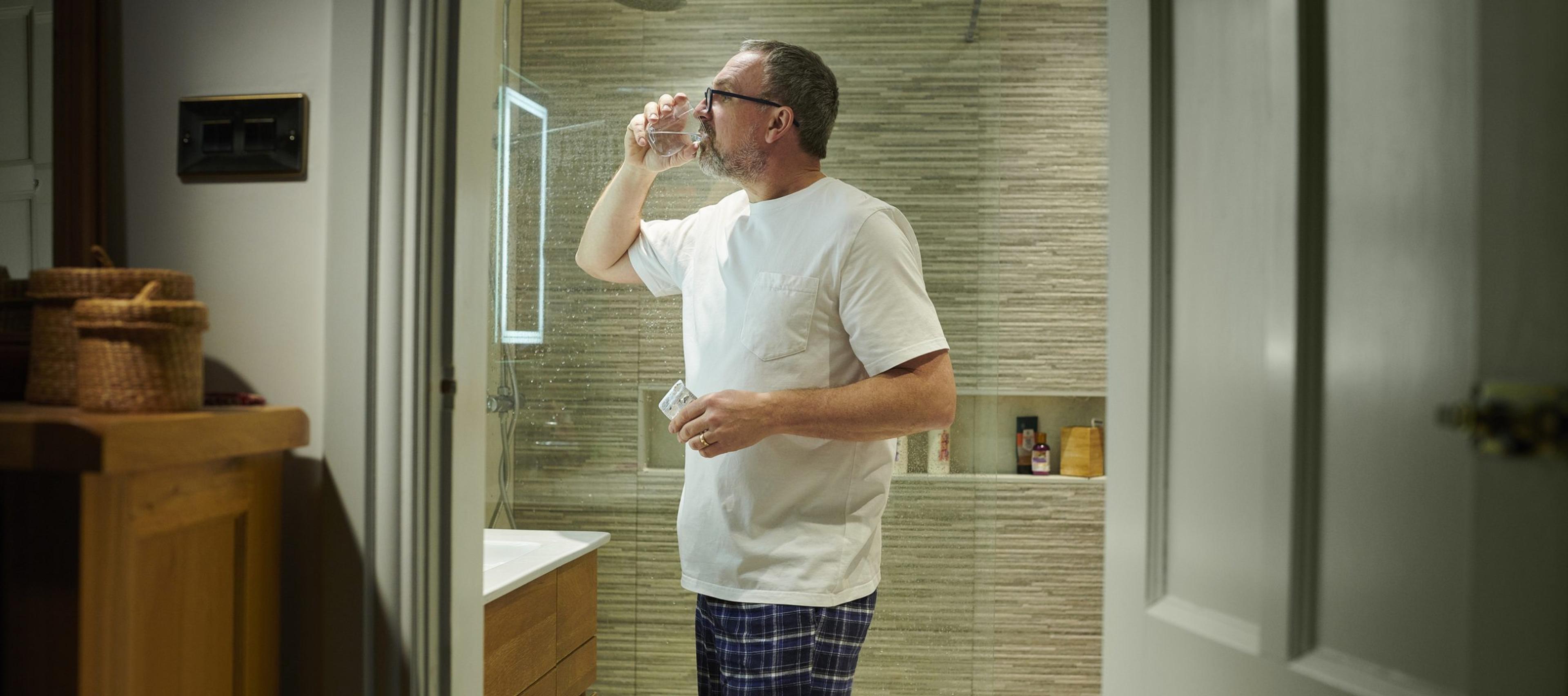 Older man drinks a glass of water in his bathroom as he does colonoscopy prep