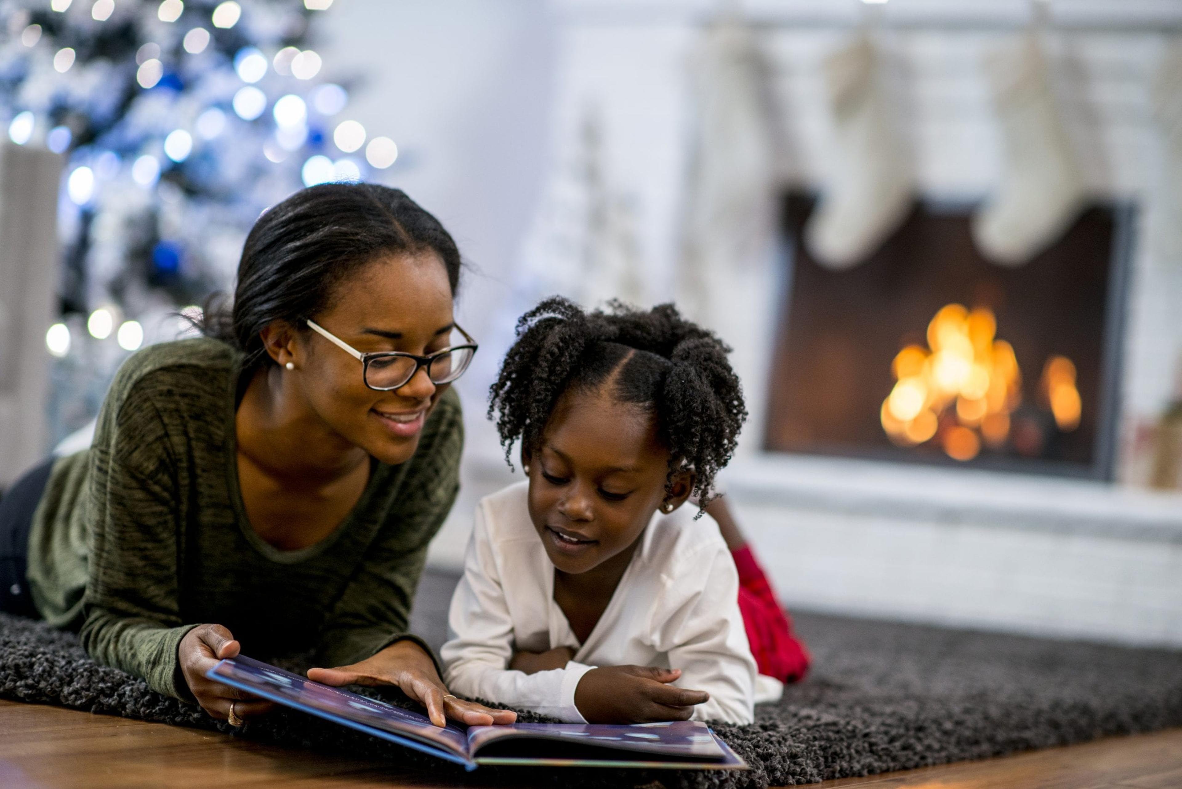 Mom and daughter read a book in front of the Christmas tree and a lit fire.