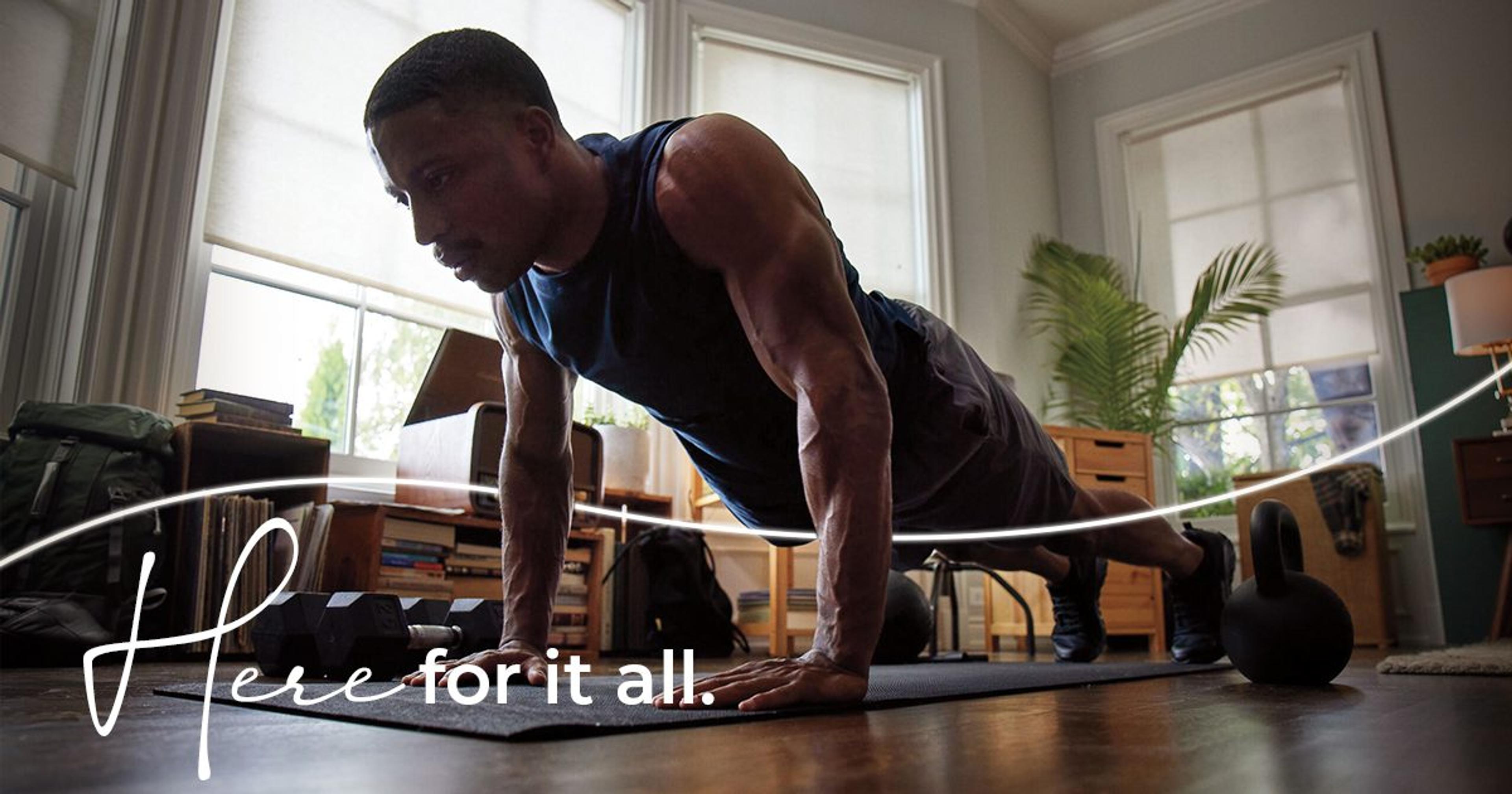 A Black man does a push up in a living room with white walls and two large windows with sunlight coming through. The text Here for it All reads across the image.
