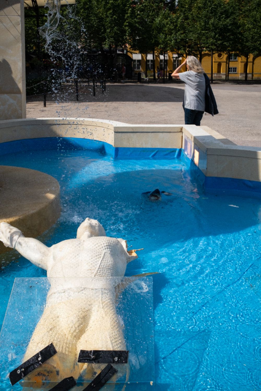 Eine umgekippte Statue liegt in einem Wasserbecken in der Stockholmer Innenstadt. Die Statue und das Becken stehen dort zur Vermarktung eines Films. Eine Frau wendet sich mit der Hand am Kopf vom Wasserbecken ab.