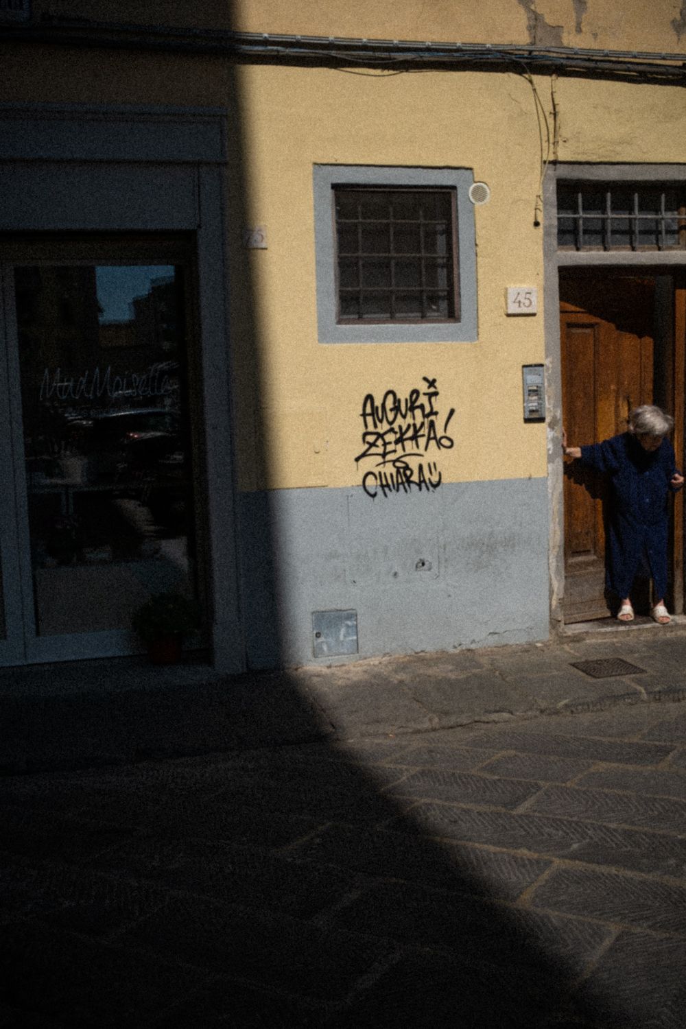 Eine ältere Frau steht in der Tür und blickt in der Altstadt von Florenz auf die Straße. Der Schatten halbiert das Bild. Sie befindet sich in der Sonne.
