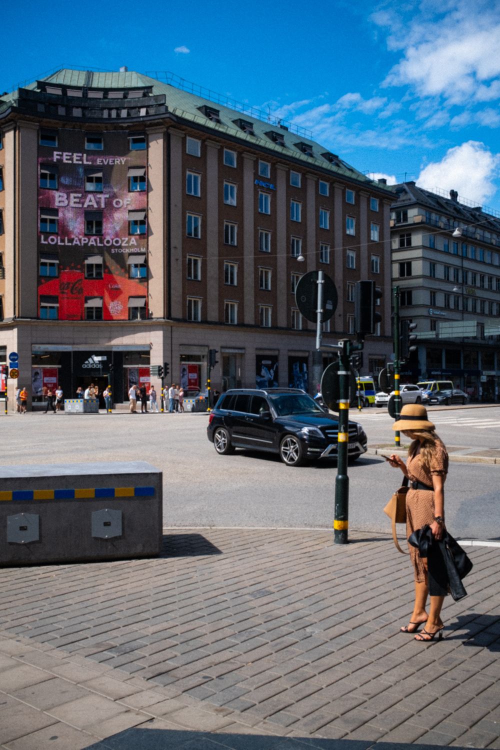 Street Photography Stockholm Frau mit Sommerhut steht an einer Stockholmer Straßenkreuzung. Sie steht allein und schaut auf ihr Smartphone