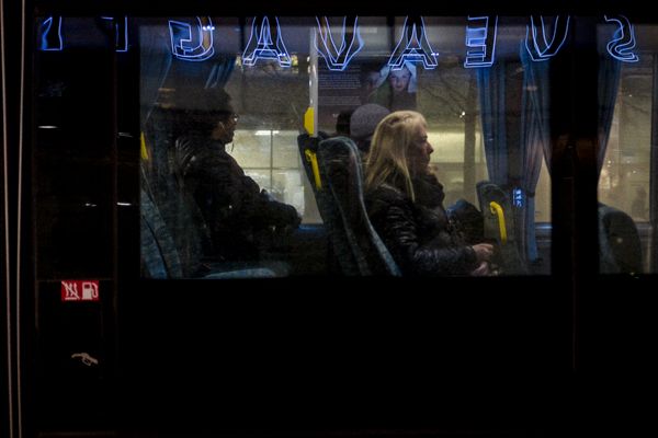 Street Photography Stockholm - Eine Frau sitzt in der Nacht in einem Bus, auf dessen Scheiben sich Leuchtreklamen spiegeln.