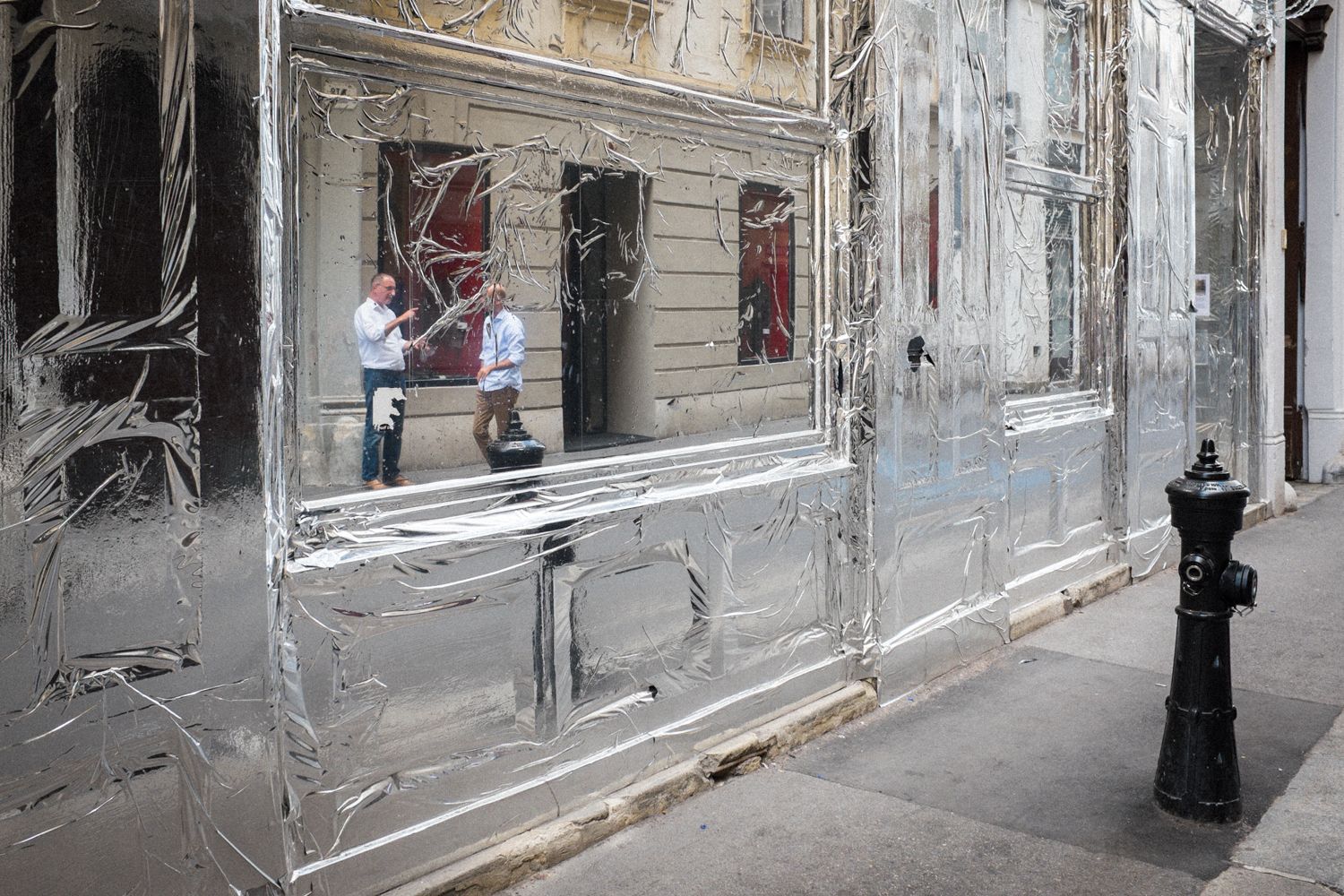 Die Fassade eines Geschäfts ist mit Silberfolie verkleidet. In der Folie spiegelt sich eine Wiener Straßenszene mit zwei sich unterhaltenden Männern auf dem Gehweg.