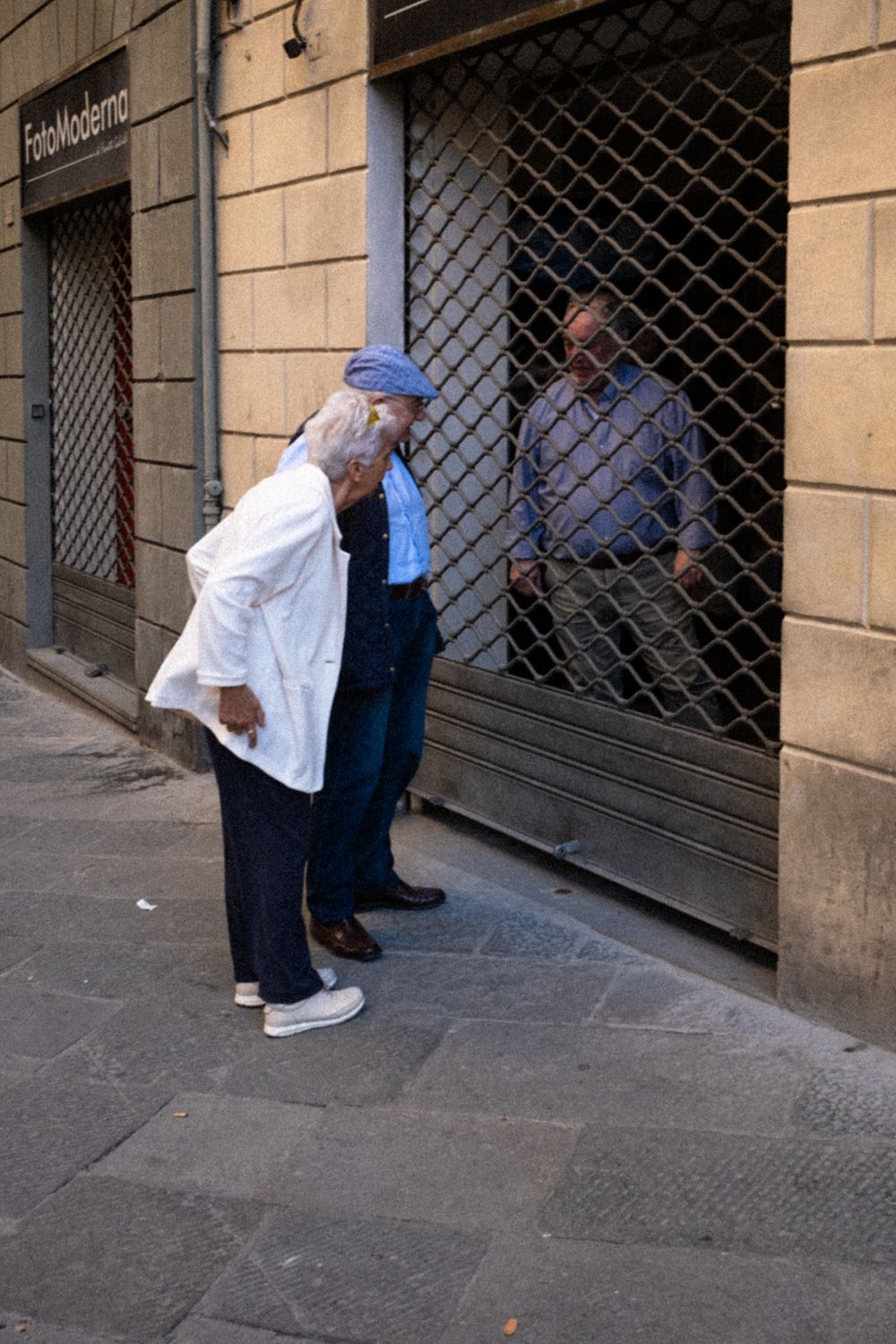 Ein Paar auf der Straße in Siena unterhält sich mit einem Mann hinter einem Gitter, das die Tür seines Geschäfts schützt.