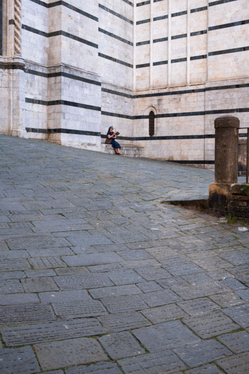 Nonne sitzt vor weißer Marmorfassade am Dom von Siena. Im Vordergrund erstreckt sich das dunkle Pflaster, typisch für die Altstadt von Siena.