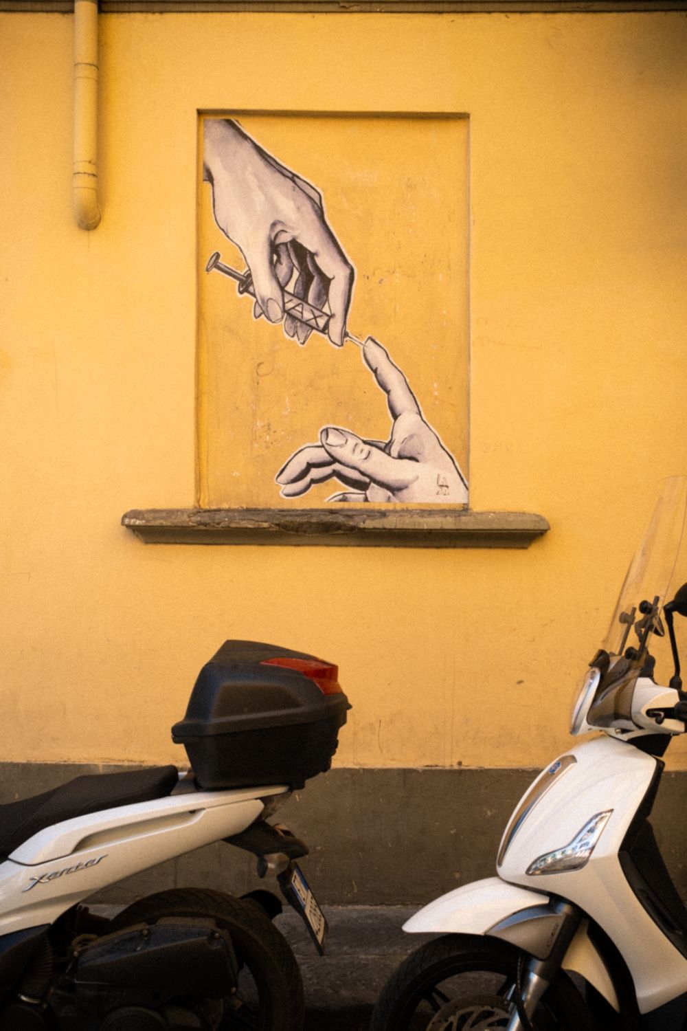 Street Art in Florenz. In einem blinden Fensterrahmen befindet sich ein Mural mit der Adaption von Gottes Hand aus der Sixtinischen Kapelle in Rom. Gott hält hier aber eine Spritze in der Hand und sticht in die andere.