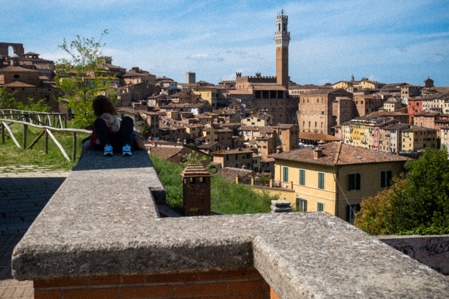 Ein Paar sitzt auf einer Mauer vor der Altstadt von Siena. Im Hintergrund eröffnet sich das Panorama auf die engen Gassen und die mittelalterlichen Gebäude.