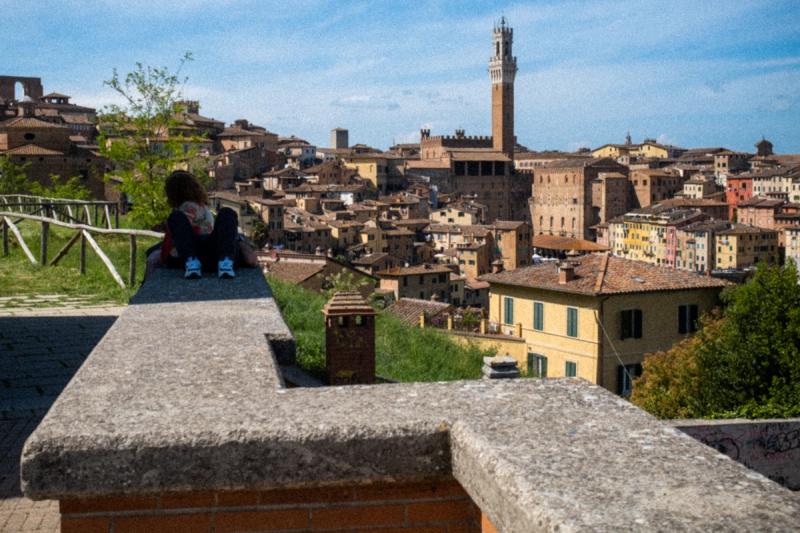 Street Photography Siena Paar liegt auf einer Mauer. Im Hintergrund eröffnet sich der Blick auf den Hügel mit der Altstadt von Siena.