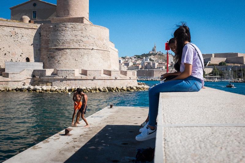 Street Photography aus Marseille. Marseille die Großstadt am Mittelmeer. Jugendliche Mädchen sitzen auf den Hafenanlagen und beobachten Jungs die im Mittelmeer baden. Auf dem Hügel im Hintergrund ist die Kirche Notre-Dame-de-la-Garde zu sehen. Sie ist das Wahrzeichen von Marseille.