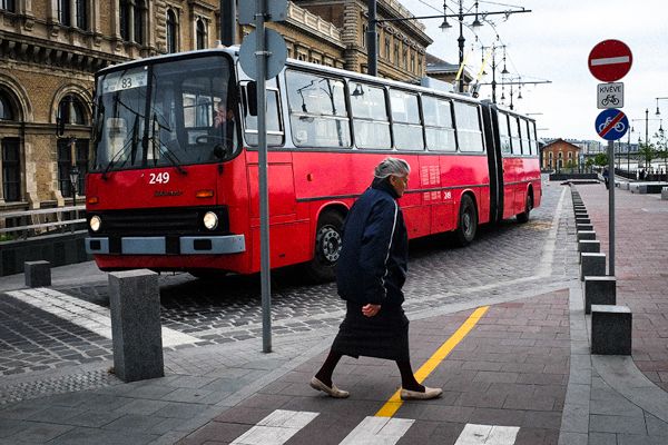 Street Photography aus Budapest, der Hauptstadt Ungarns. Eine Rentnerin überquert die Straße. Ein Ikarus Bus lässt sie passieren. Der Ikarus Bus wird in Ungarn hergestellt und ist in vielen ehemals kommunistischen Ländern weit verbreitet.