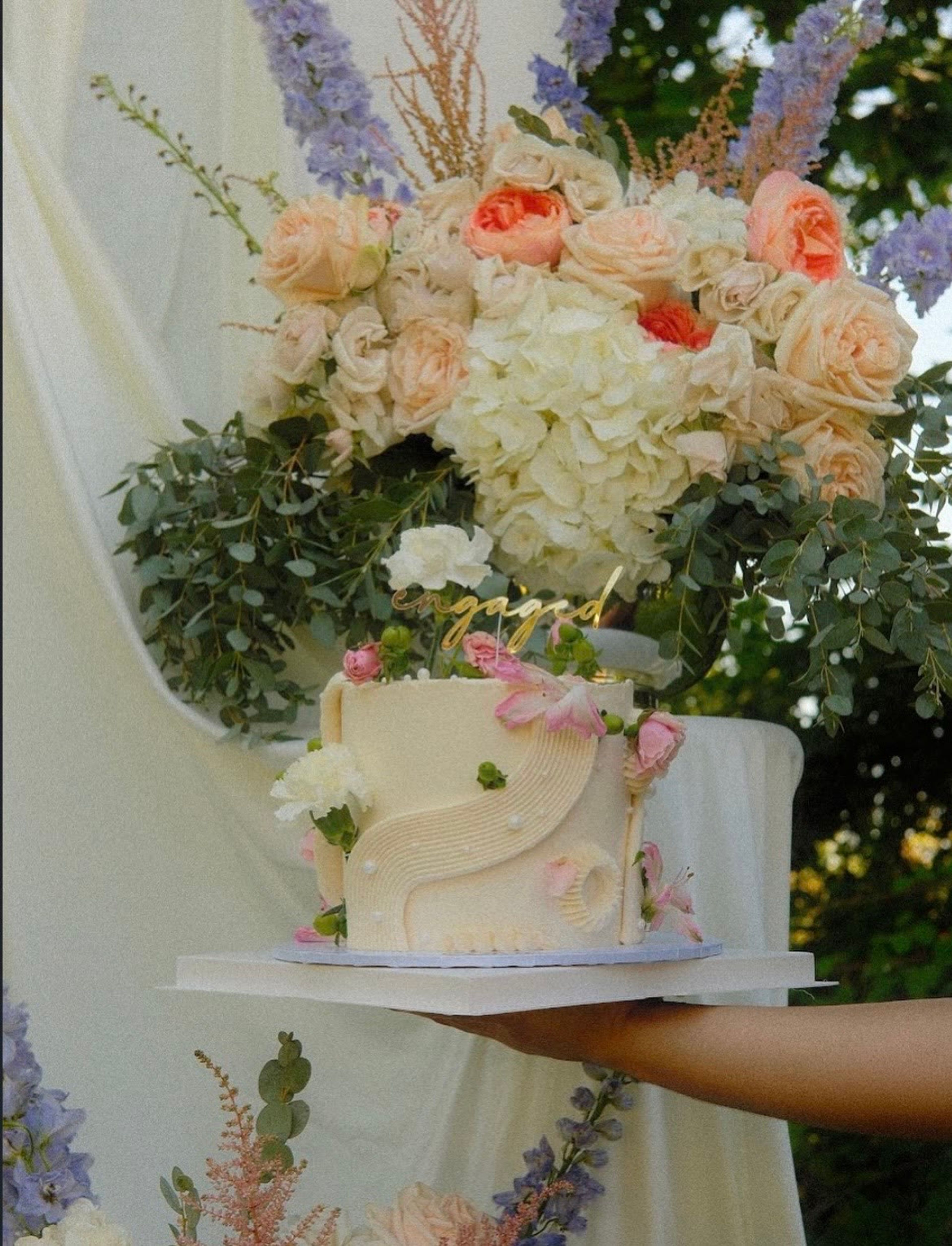 A tiered cake held in front of cream drapery and flowers
