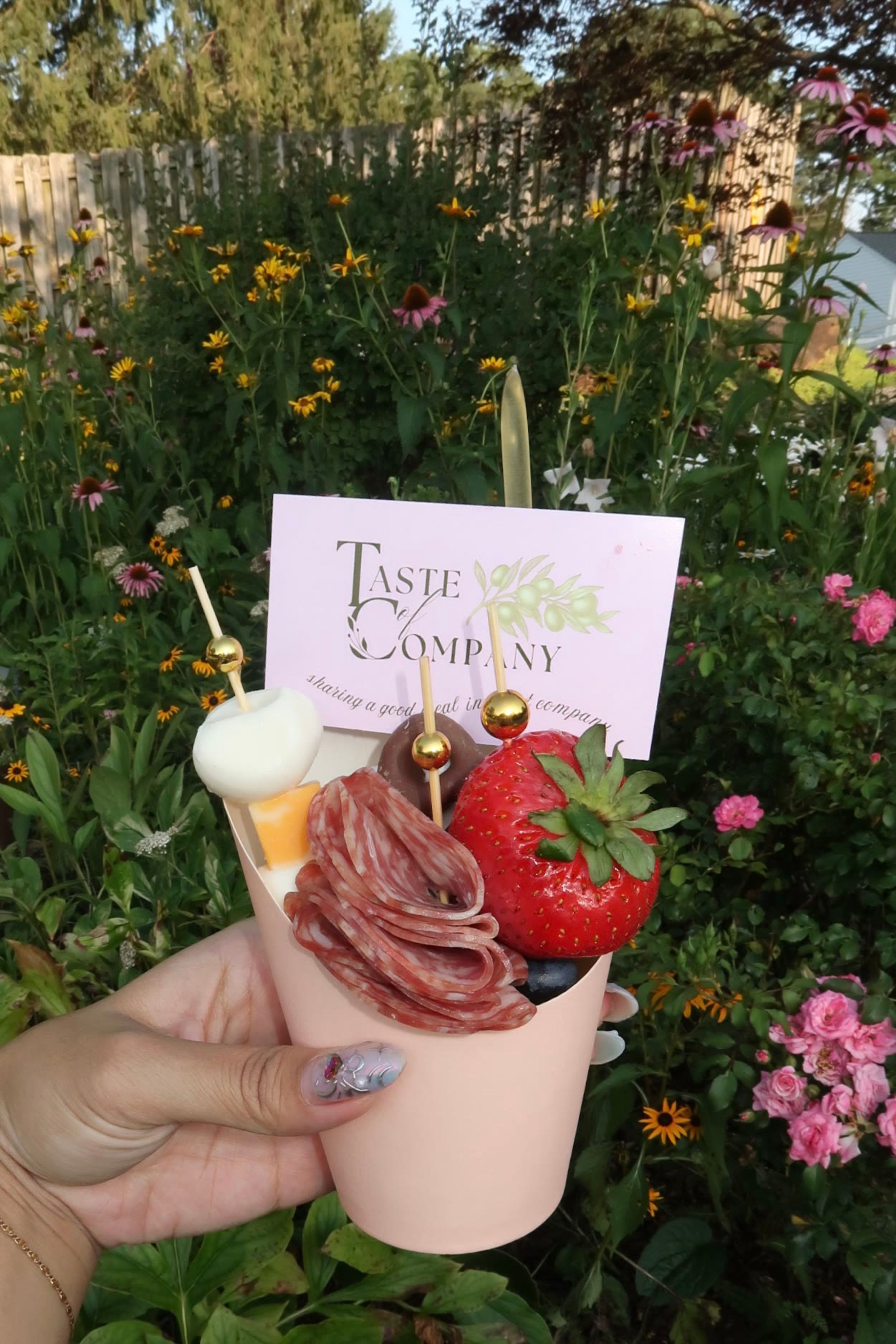 A charcuterie cup held up in front of wildflowers, with a Taste of Company card