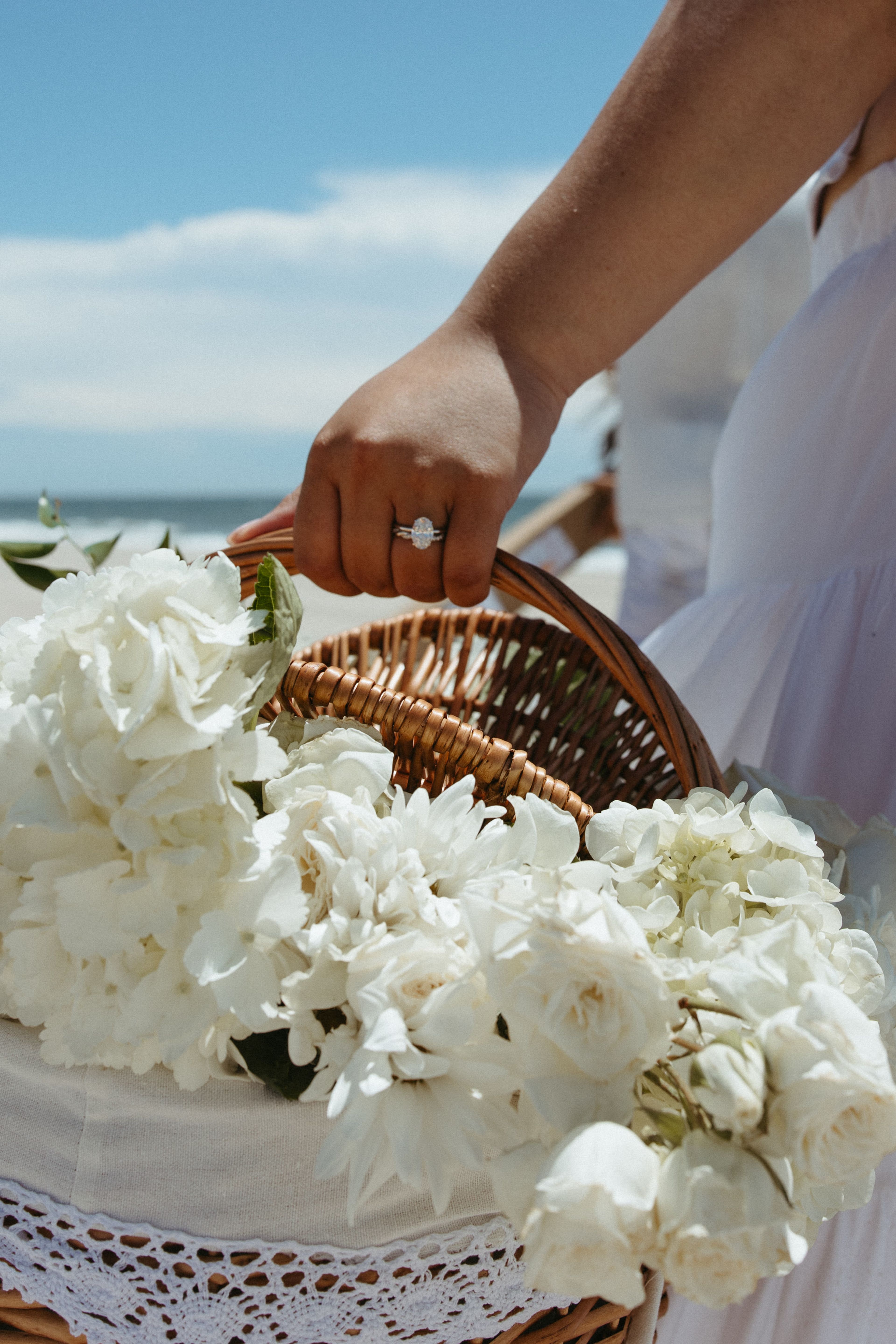 A hand wearing an engagement ring resting on a basket of white flowers
