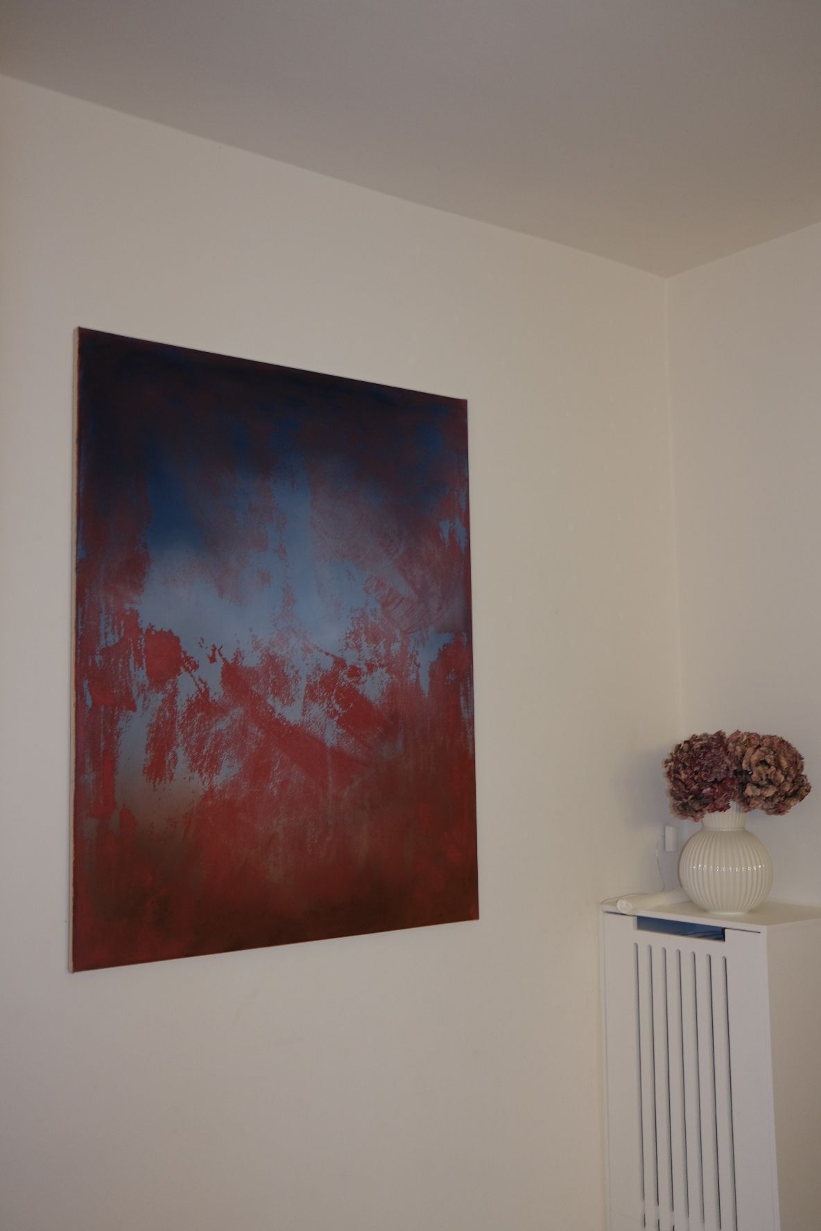 Abstract painting in portrait format. A deep navy top fades into a hazy pale blue, which is broken up from below by rough, scraped strokes of rust red that take over the lower half. From an angle.