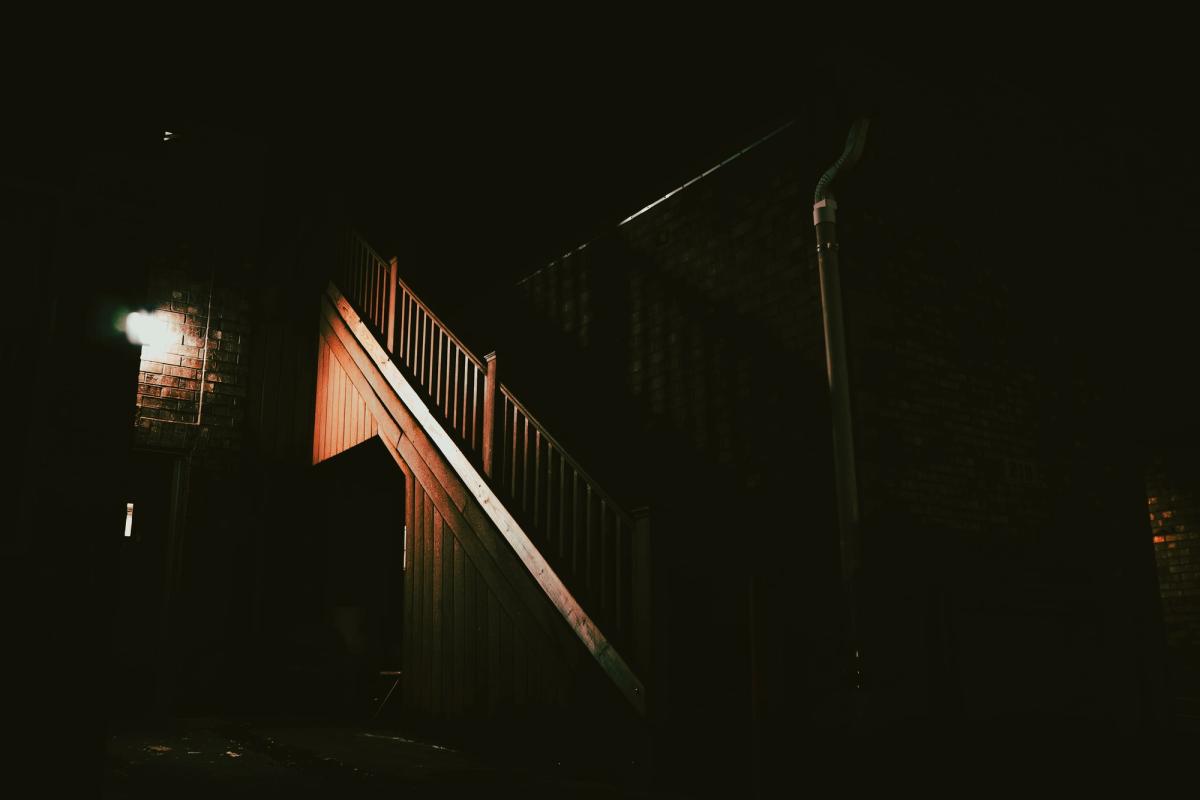 An outdoor wooden staircase beside a brick building, lit by one wall lamp at night.