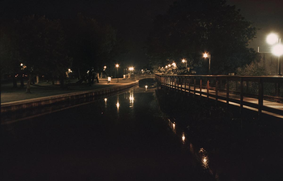 A canal at night between a lamplit park path and a railed footbridge, the lights doubled in the water.