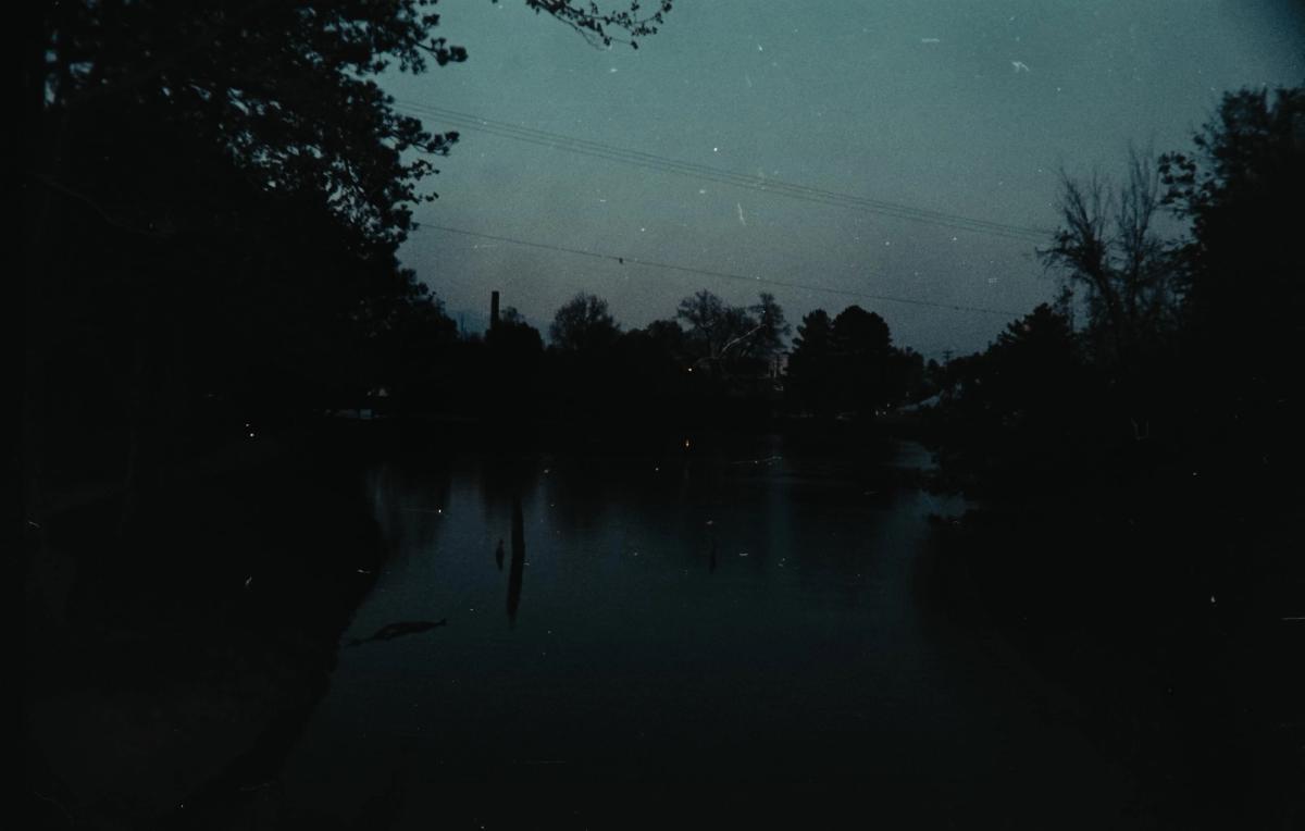 A still river at dusk between dark trees, power lines crossing a blue-gray sky.