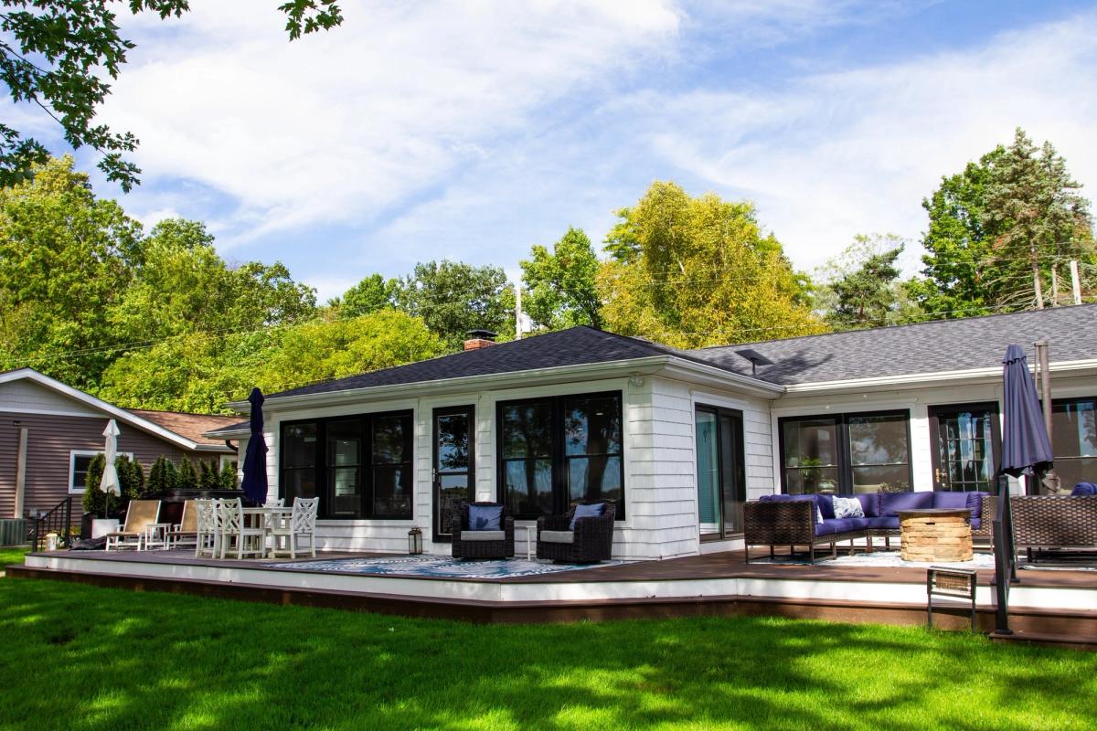A white single-story lakeside house with a dark shingled roof, a wide deck and a green lawn.