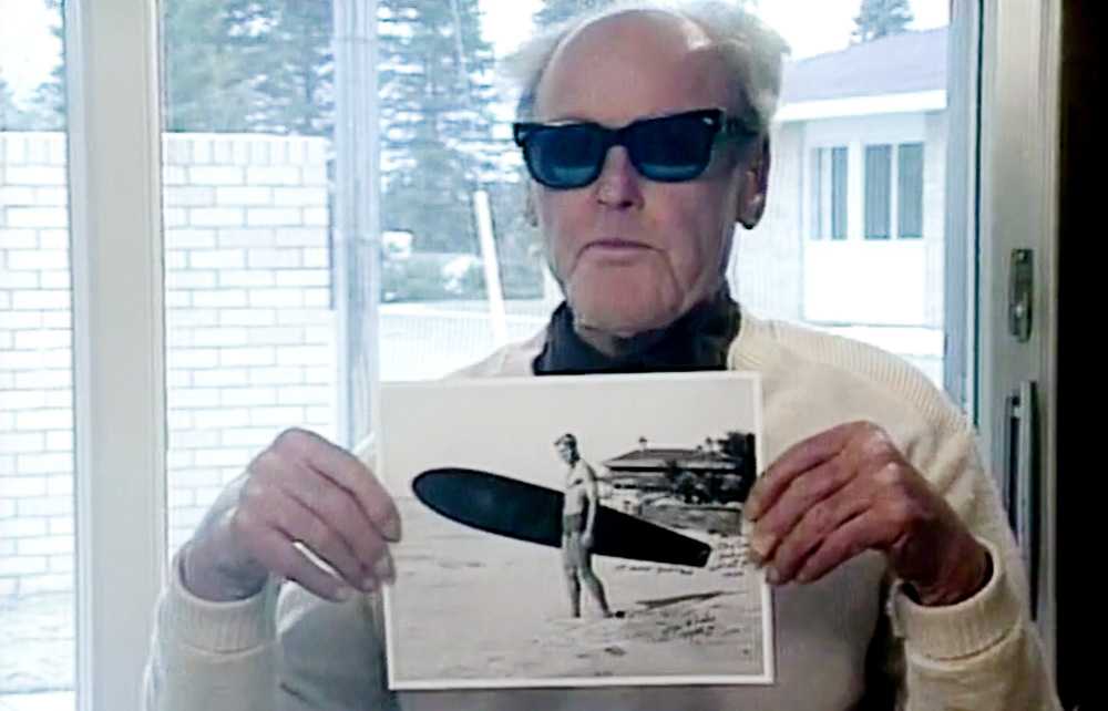 tom blake holding a photo of himself with a surfboard