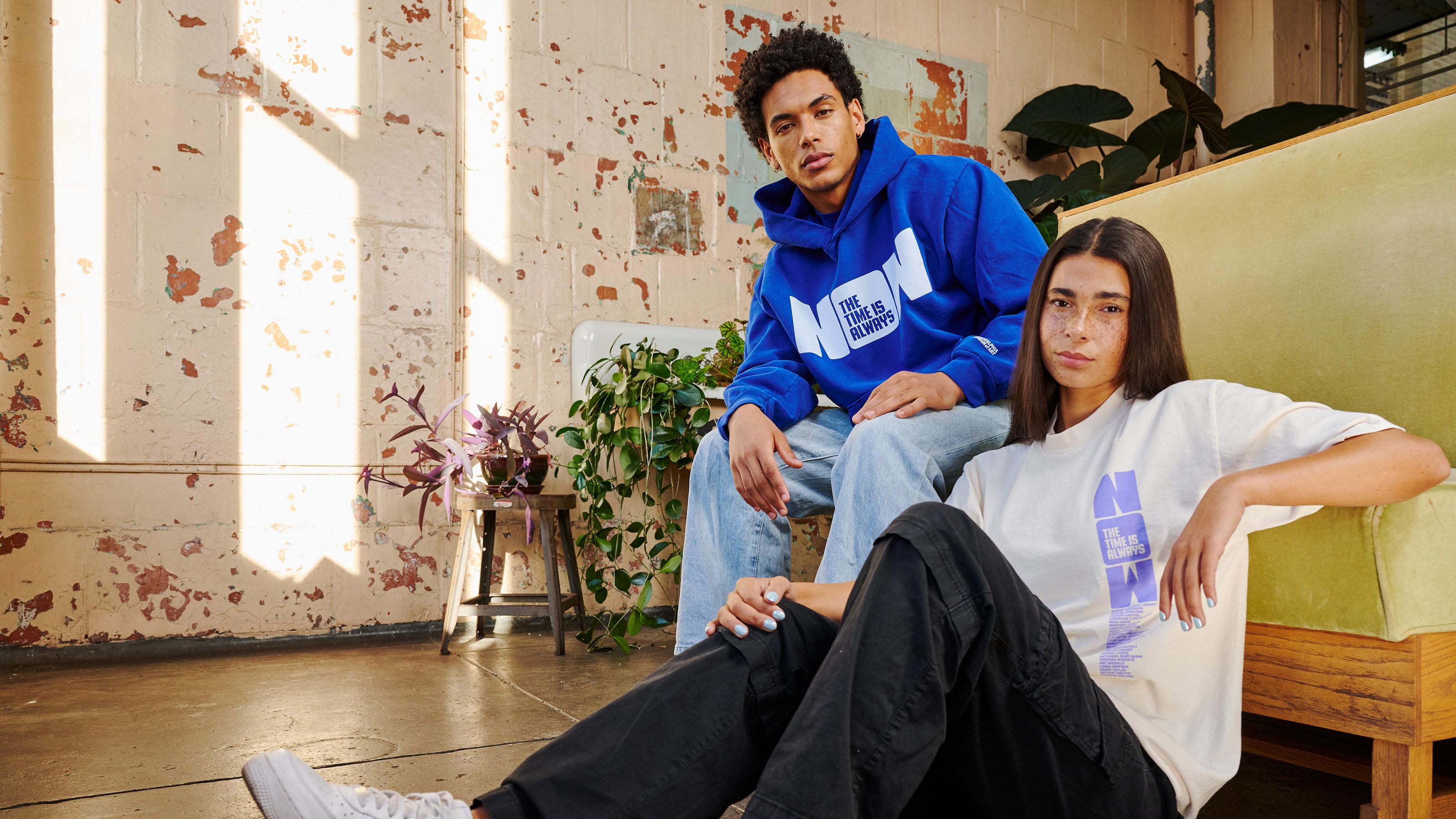 Two young adults wearing a sweartshirt and t-shirt that has the words THE TIME IS ALWAYS NOW printed on it.
