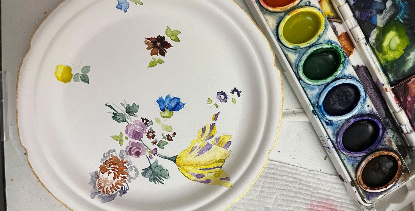 Paint palette on a table next to a white plate with painted flowers