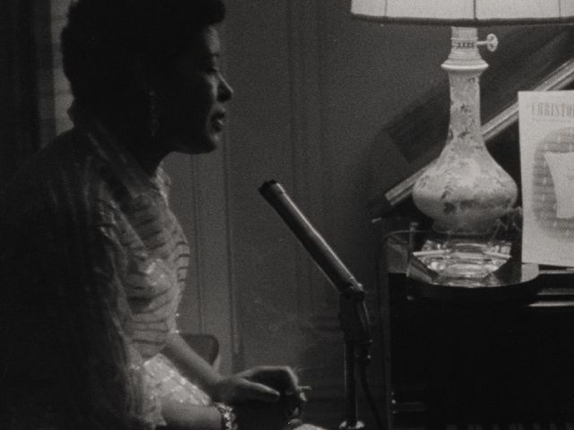 A close up of Billie Holiday sitting in a home, singing in front of microphone.