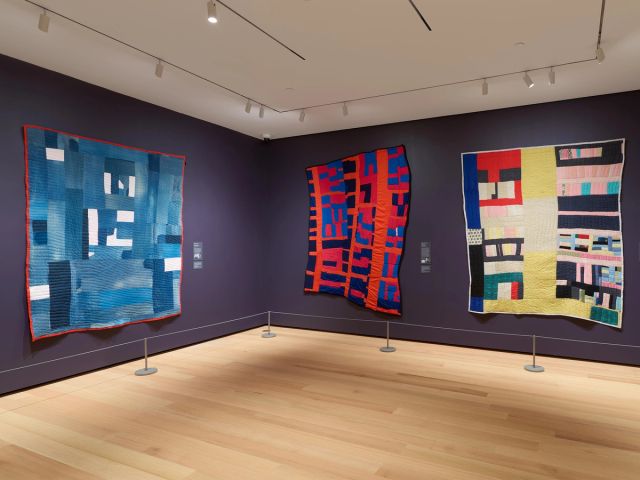 Installation view of the Gee&apos;s Bend Gallery which shows three quilts hung on the wall.