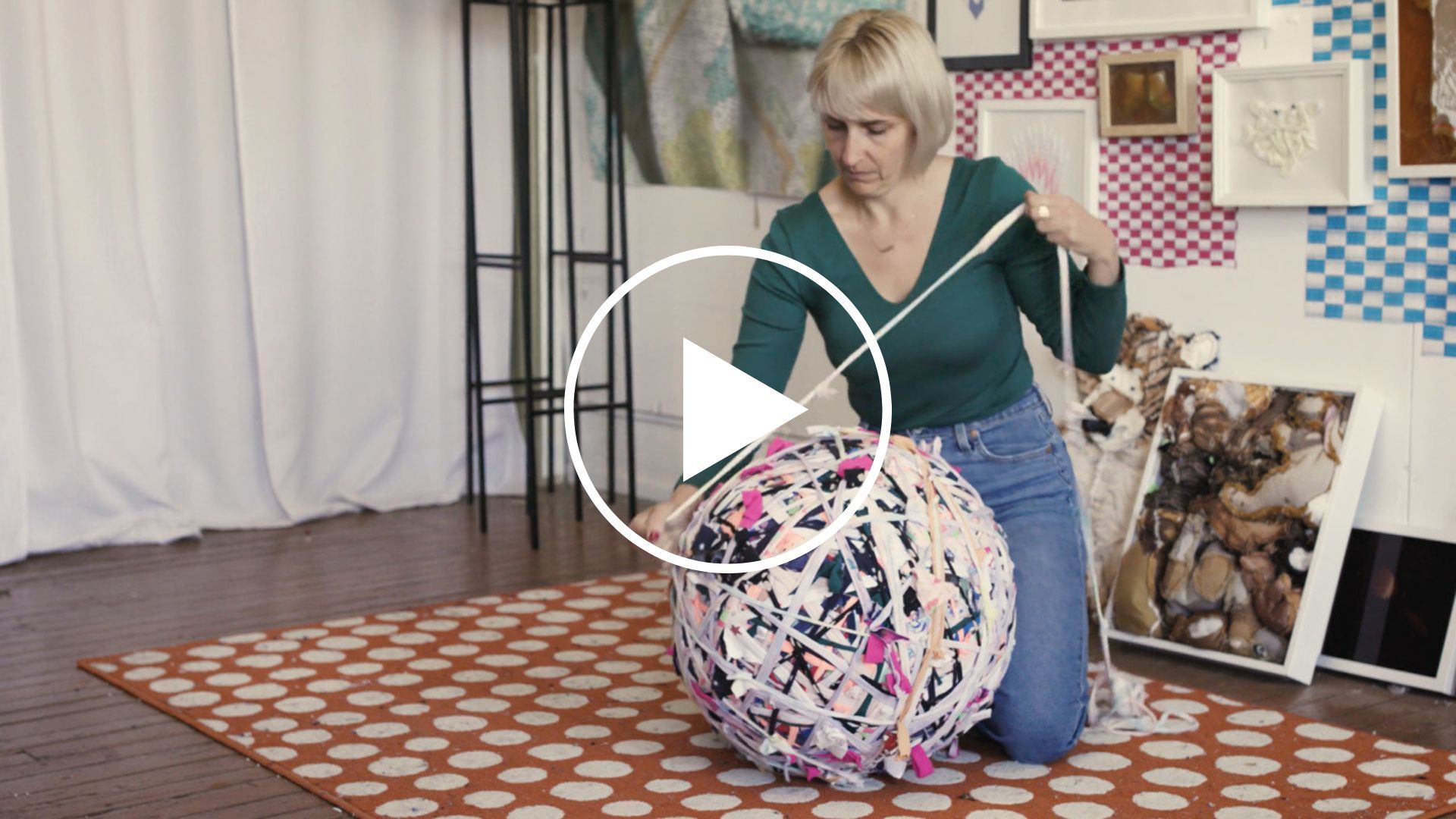 Artist with pale skin and light hair wearing a dark green top and jeans kneeling with a large ball of intertwined fabric.