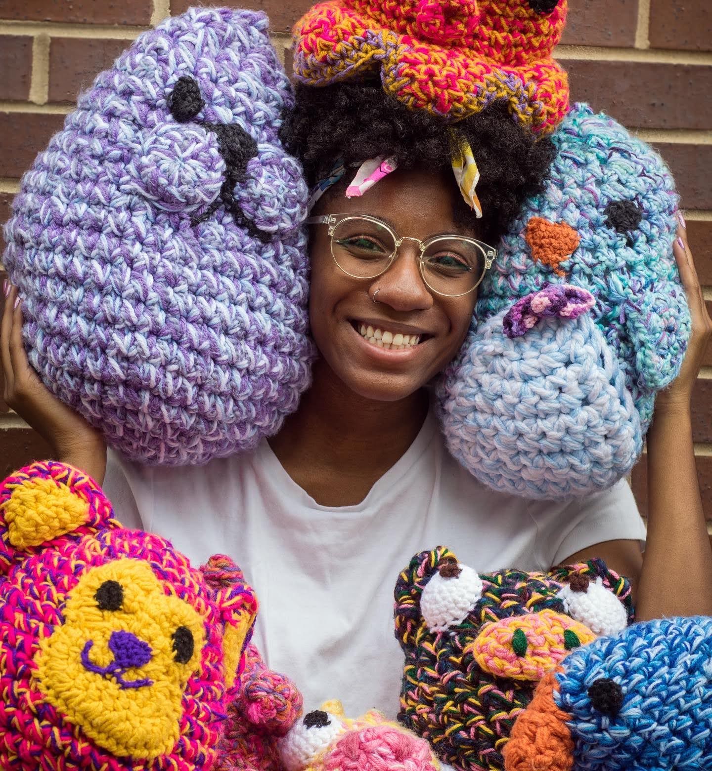 Black woman wearing a whilte shirt surrounded by colorful crochet stuffed animals.