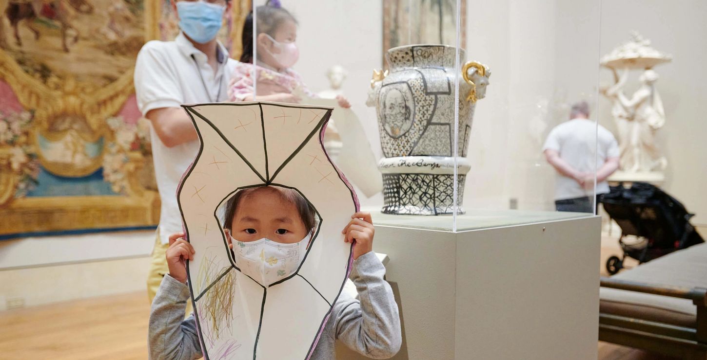 A child holding up their drawing of a nearby vase while their parent smiles in the background.