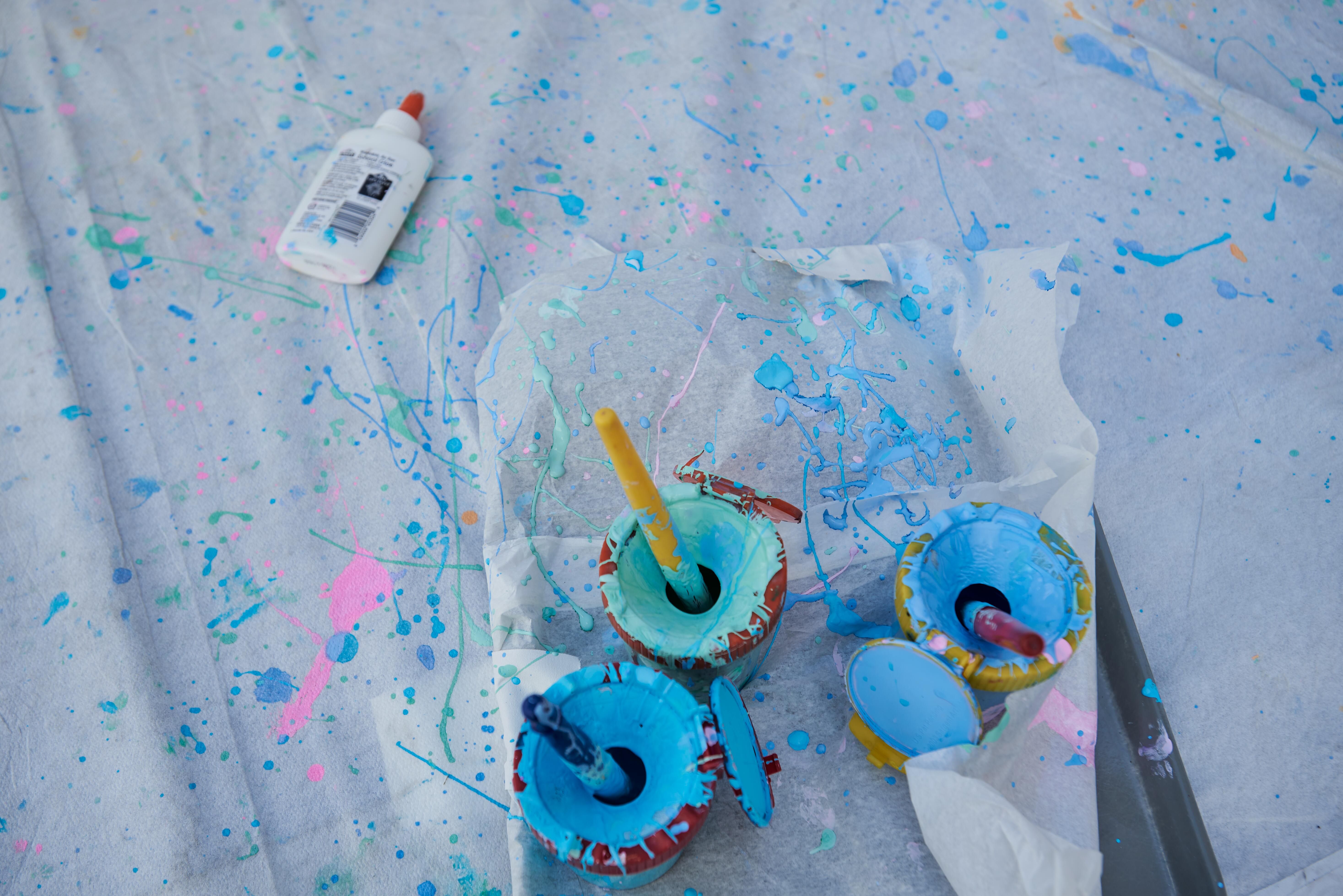 Buckets of blue and green paint on a paint splattered cloth with a bottle of glue in the upper left corner.