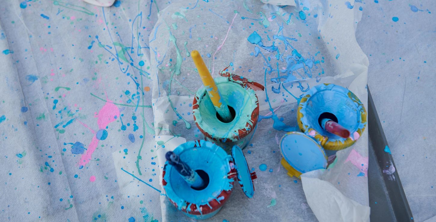 Buckets of blue and green paint on a paint splattered cloth with a bottle of glue in the upper left corner.