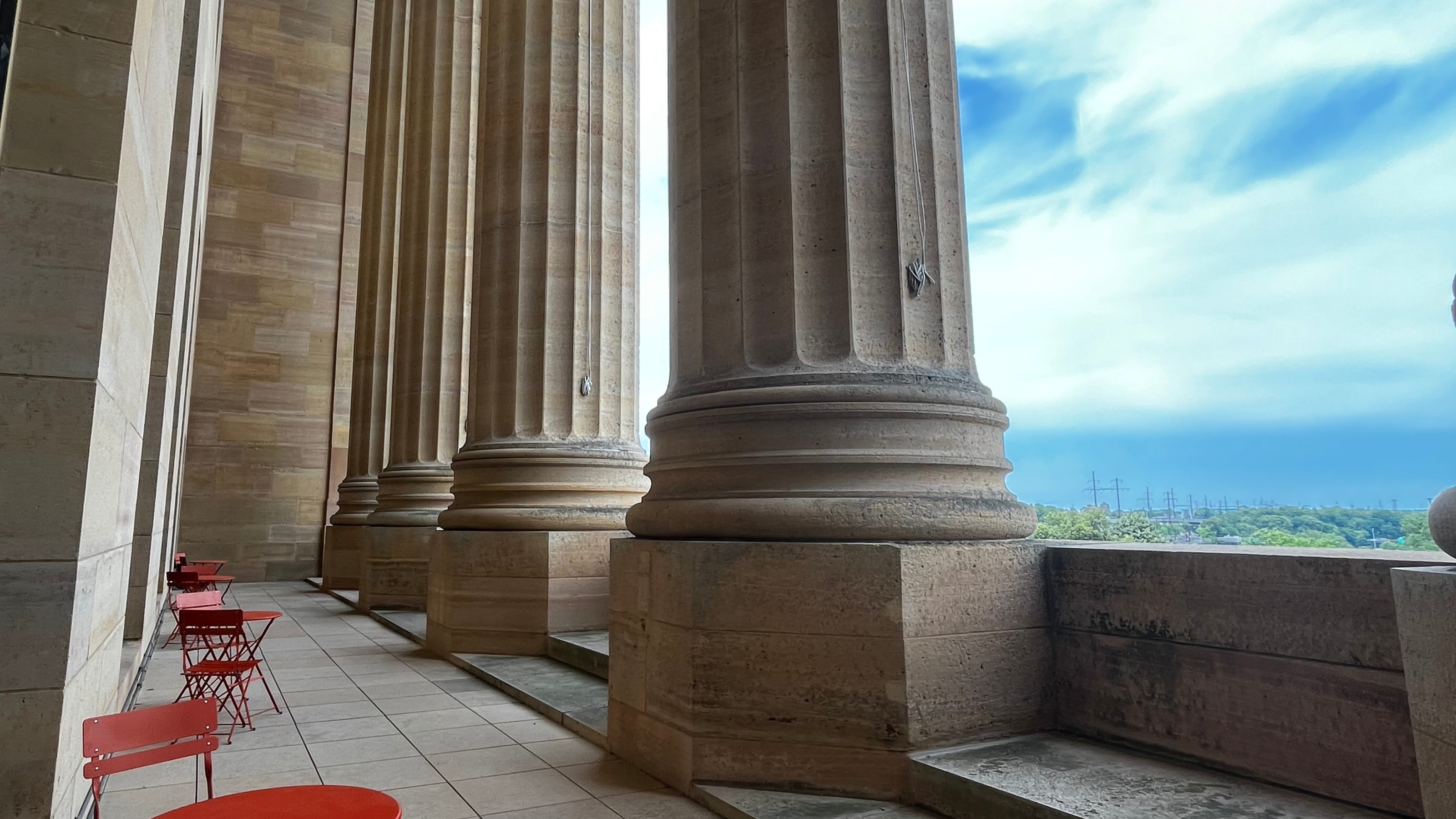 Image of a narrow terrace with red bistro tables and large columns.