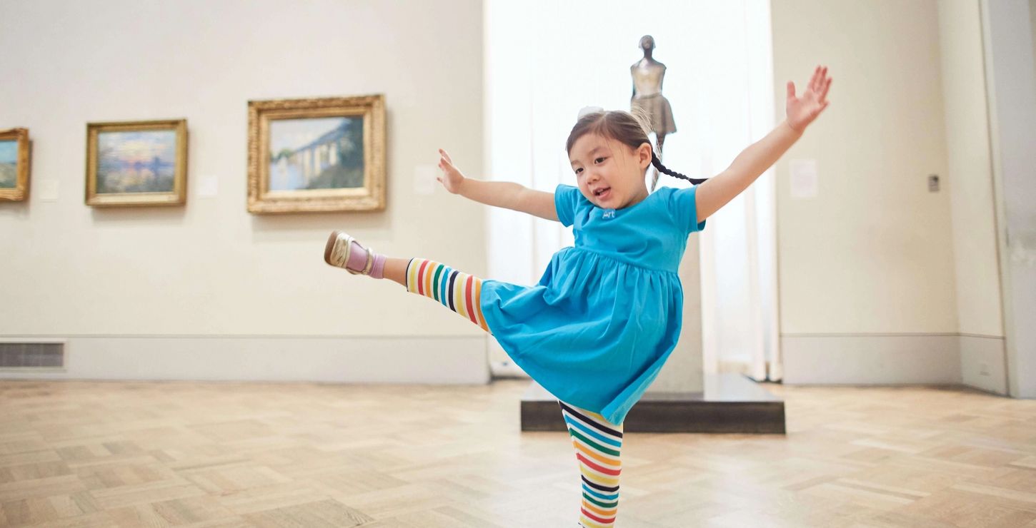 A child in a blue dress dancing in the galleries in front of the sculpture Little Dancer, Aged Fourteen by Edgar Degas